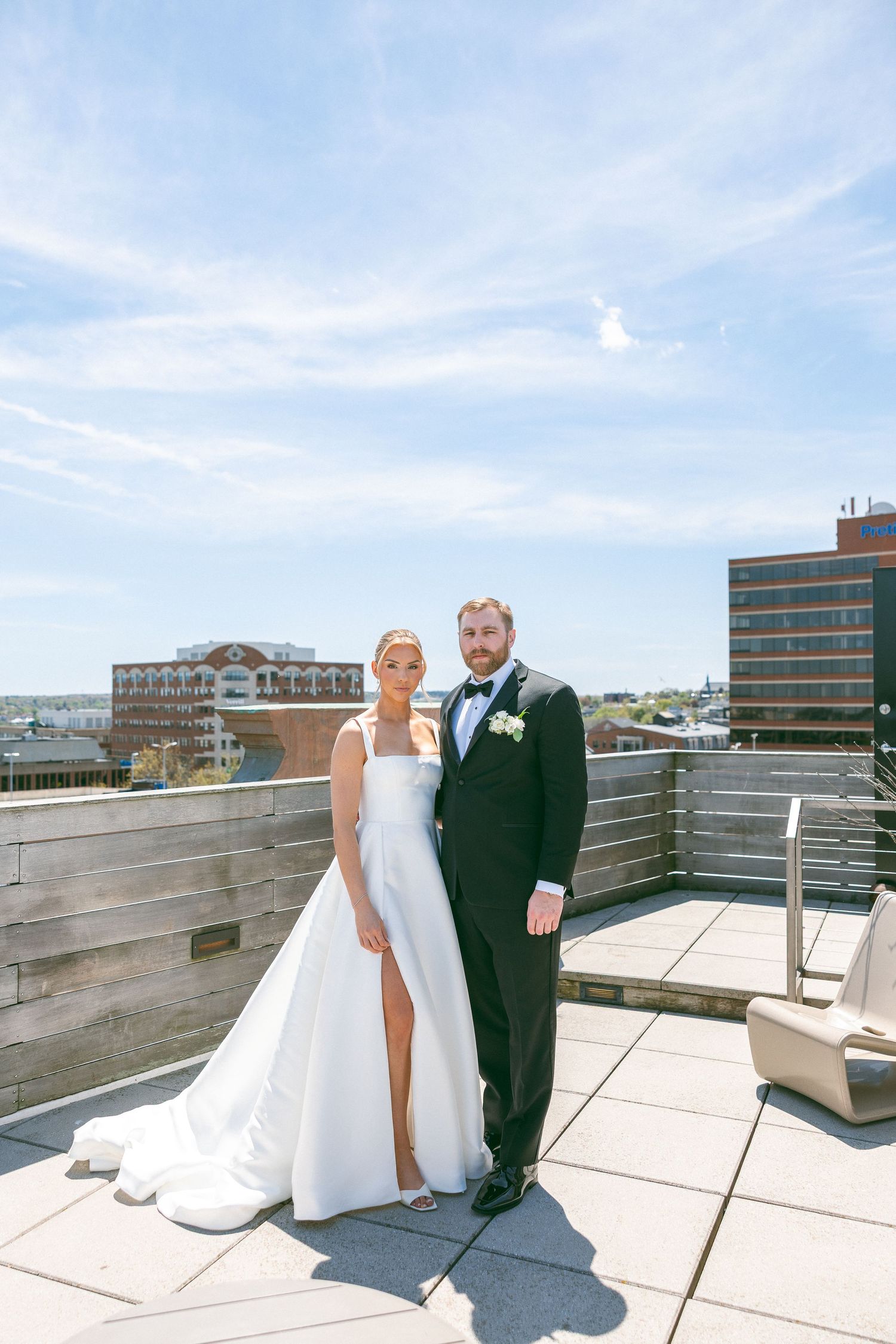 Bride and groom pose on rooftop terrace in wedding attire with city skyline backdrop on sunny day.
