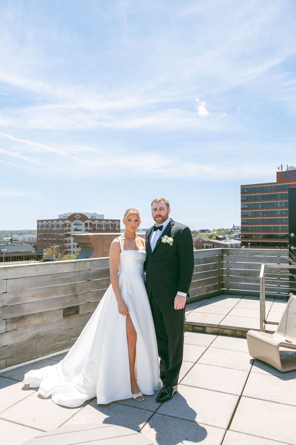 Bride and groom pose on rooftop terrace in wedding attire with city skyline backdrop on sunny day.