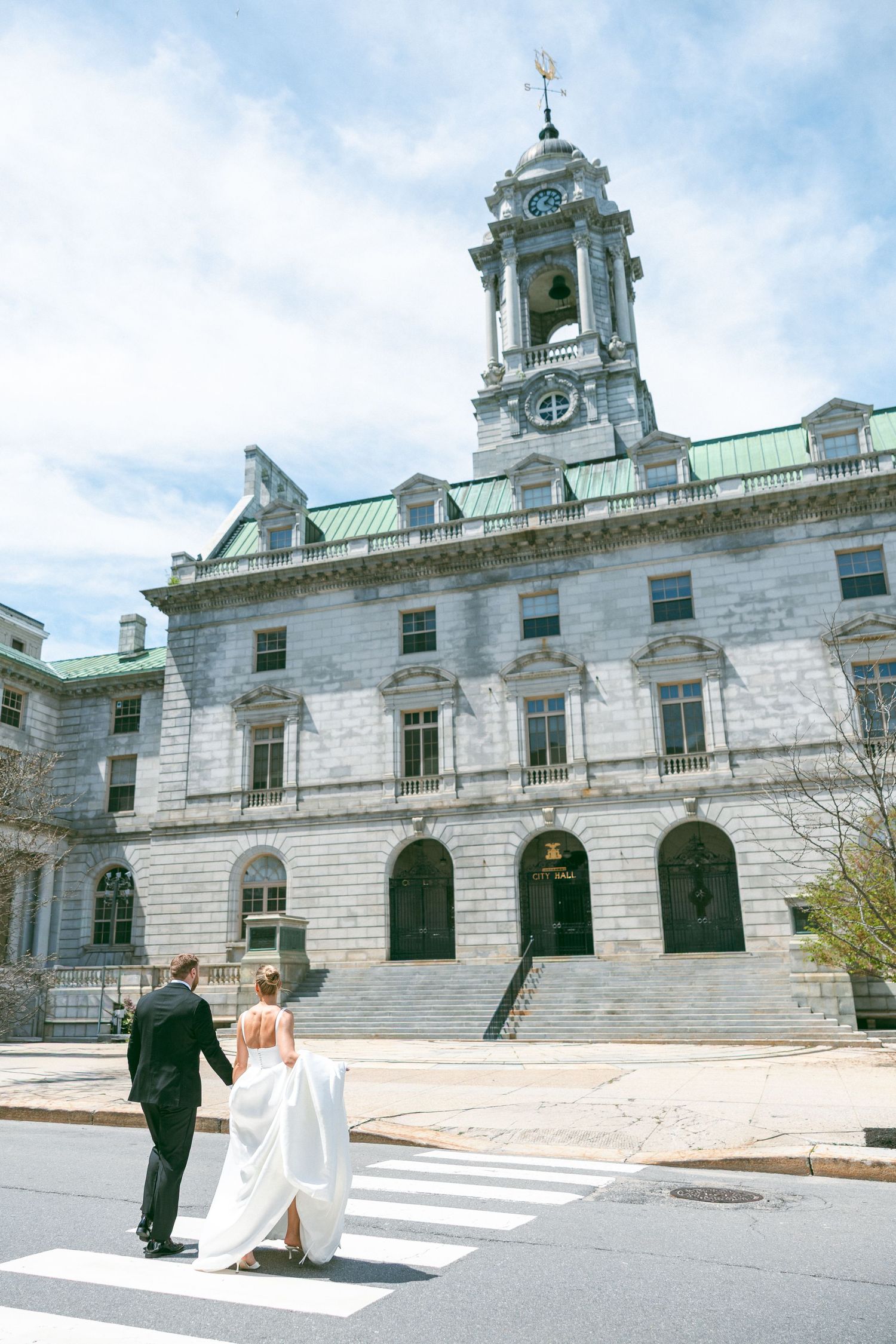 Bride and groom cross a zebra crossing hand in hand before a grand stone building with a clock tower.