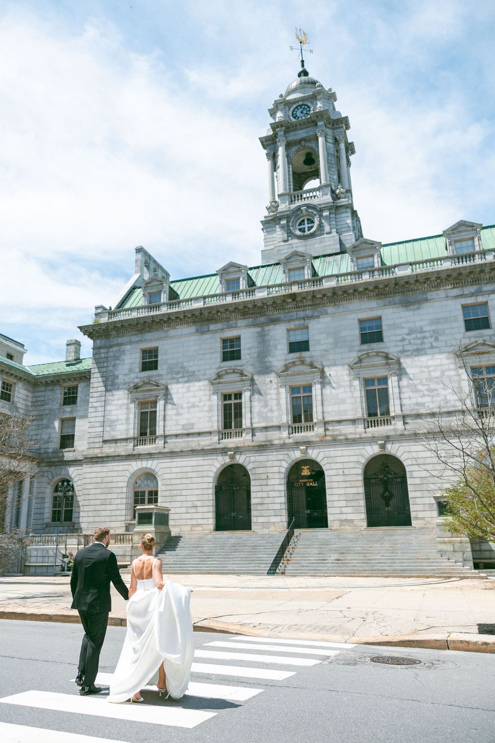 Bride and groom cross a zebra crossing hand in hand before a grand stone building with a clock tower.