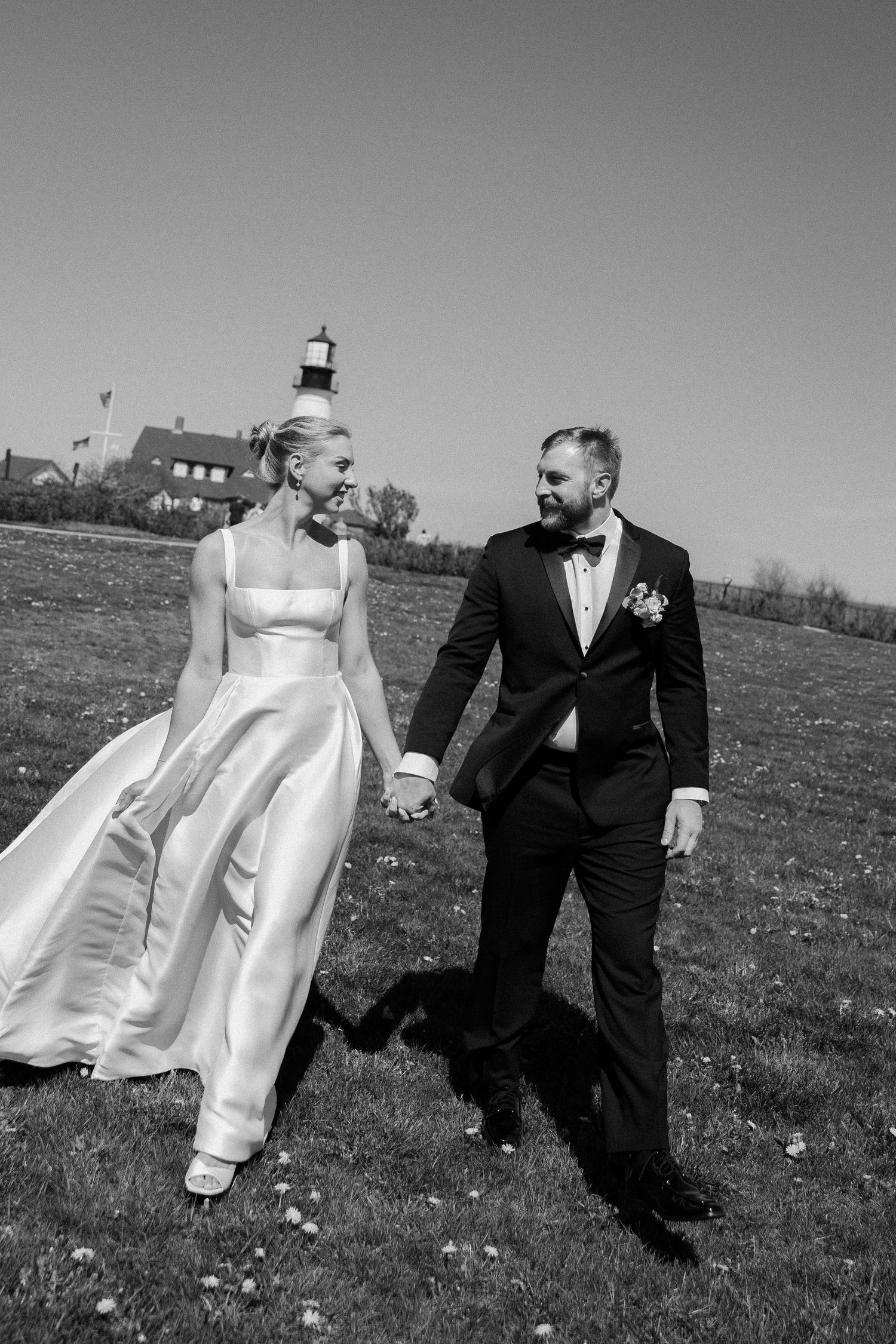 Black and white photo of a bride and groom holding hands, walking on grass with a lighthouse in the background.