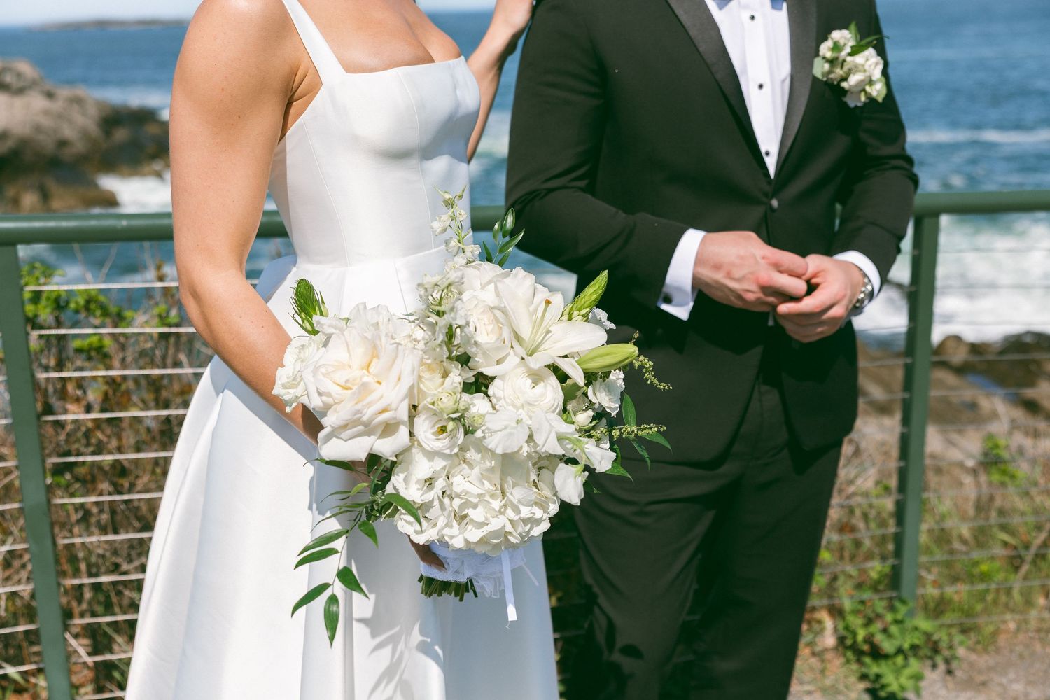Bride in white gown holding white peony bouquet stands with groom in black tuxedo by ocean railing.