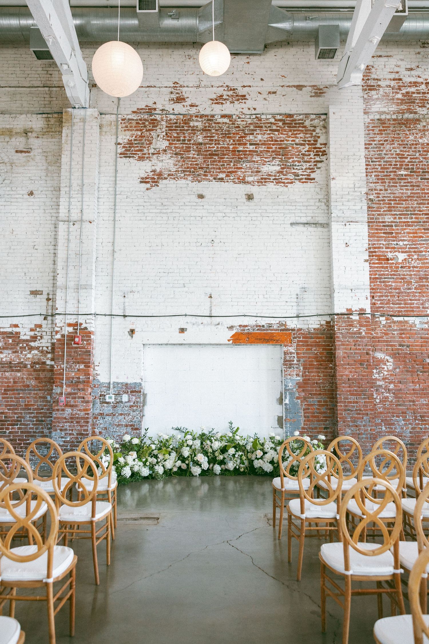 Industrial wedding venue with brick walls, white floral arch, gold chairs, and globe pendant lights.