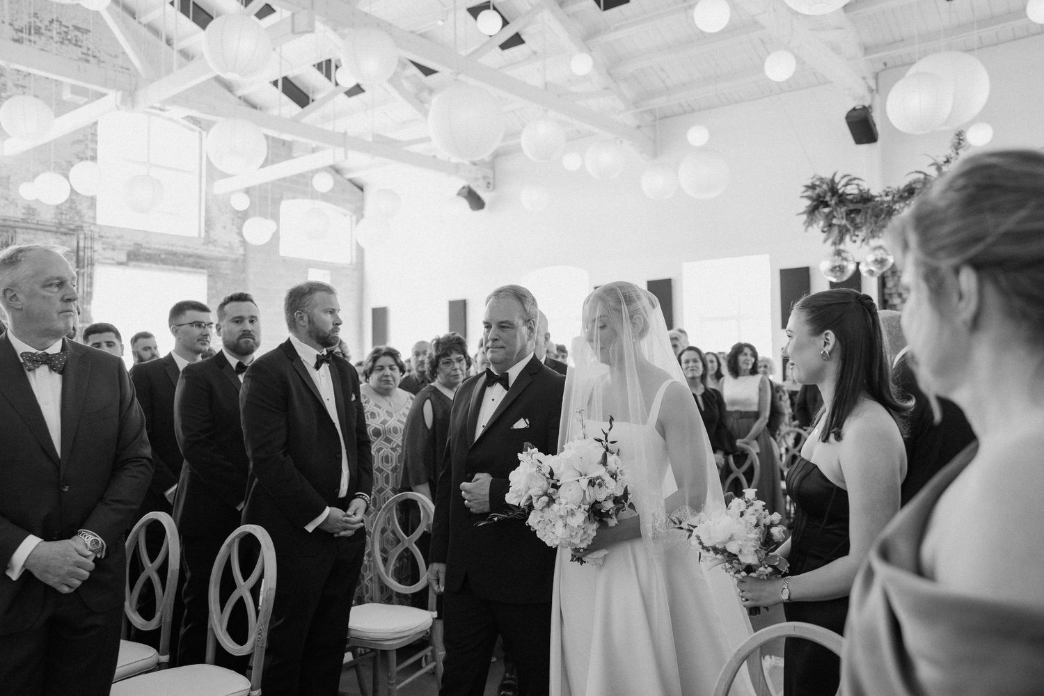Black and white photo of a bride walking down the aisle with her father during an elegant indoor wedding ceremony.
