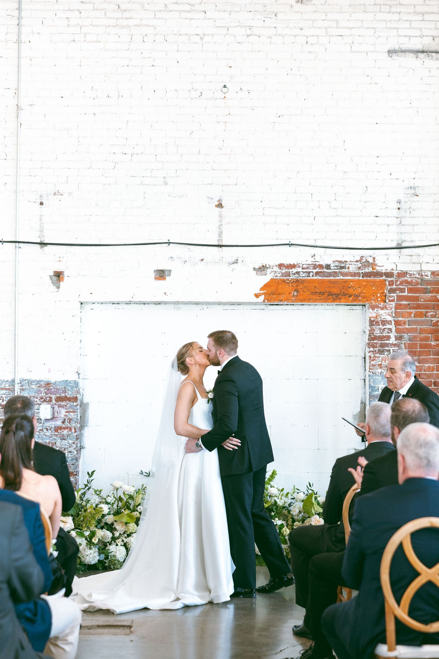Bride and groom share first kiss at industrial chic wedding ceremony with white brick walls and floral arrangements.