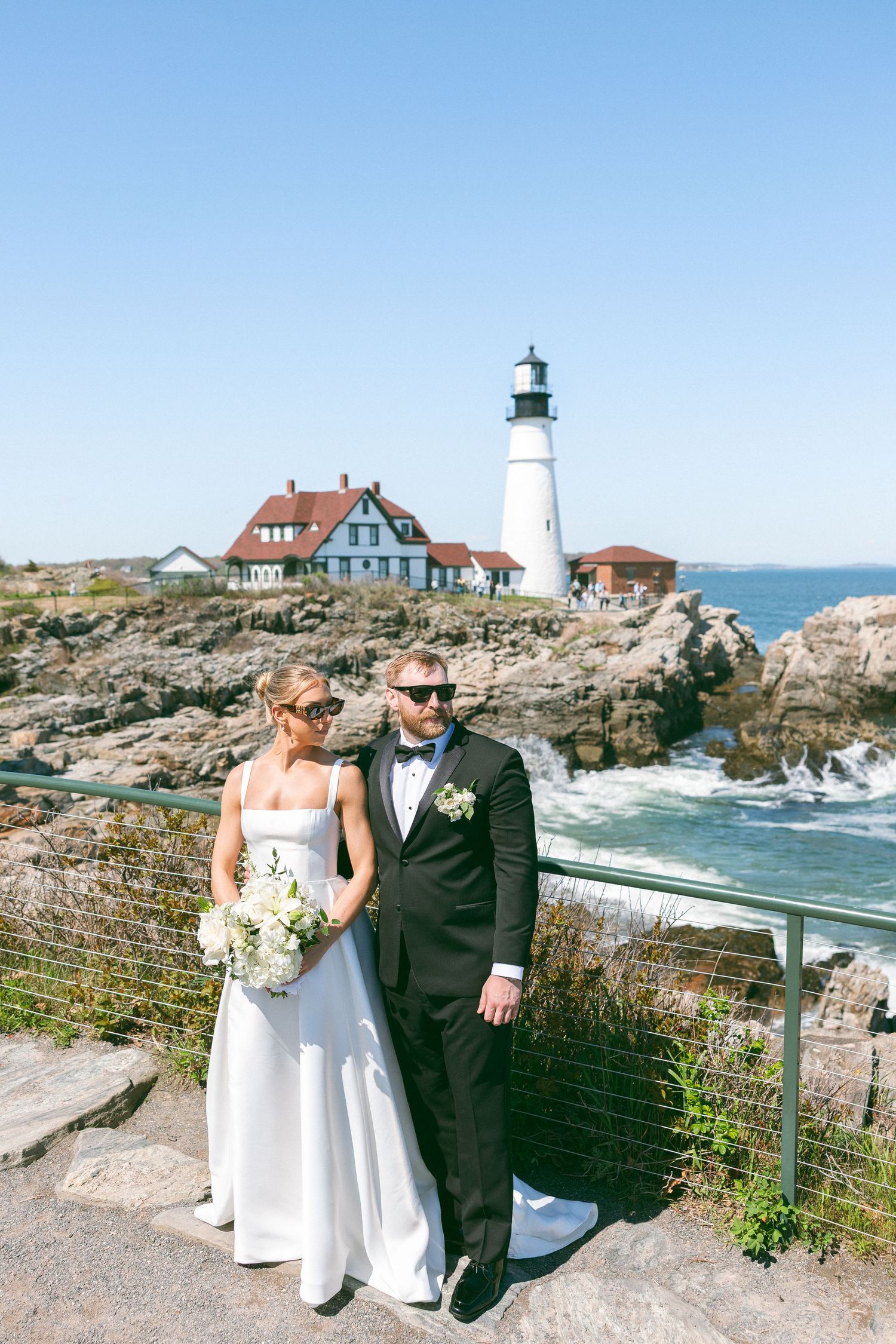 Bride and groom pose by rocky Maine coastline with Portland Head lighthouse and red-roofed buildings under clear blue sky.
