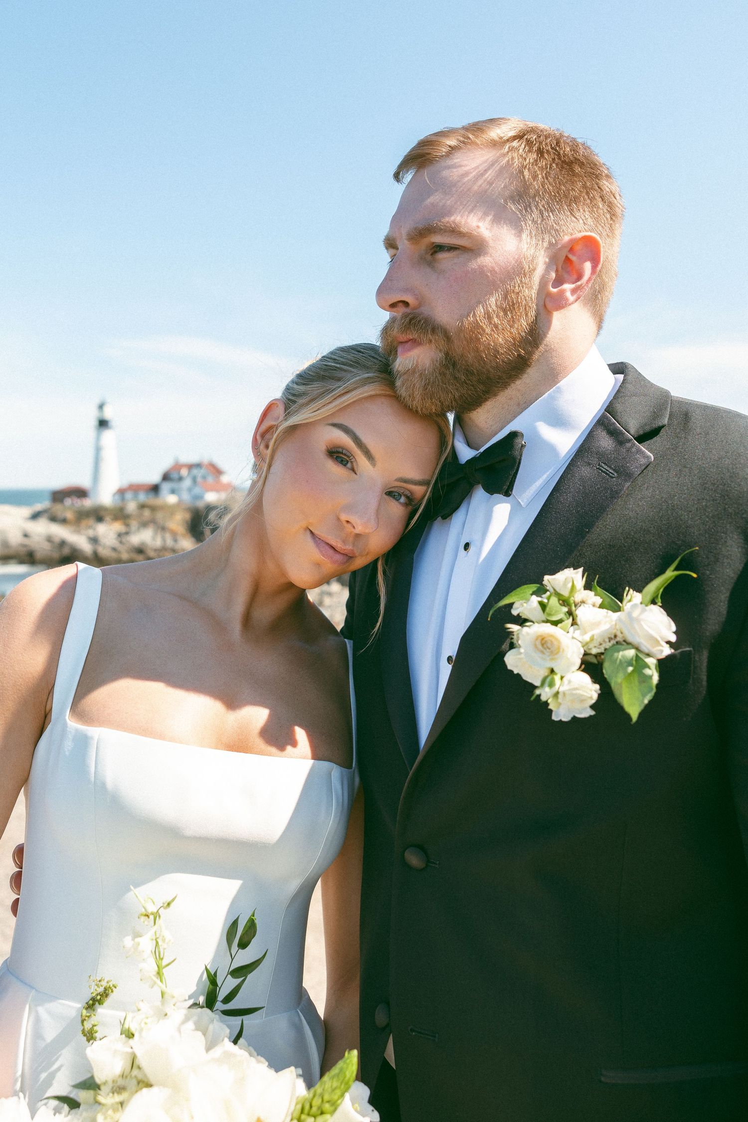 Bride and groom pose near lighthouse, she leans on his shoulder holding white bouquet, he wears black tuxedo with boutonniere.