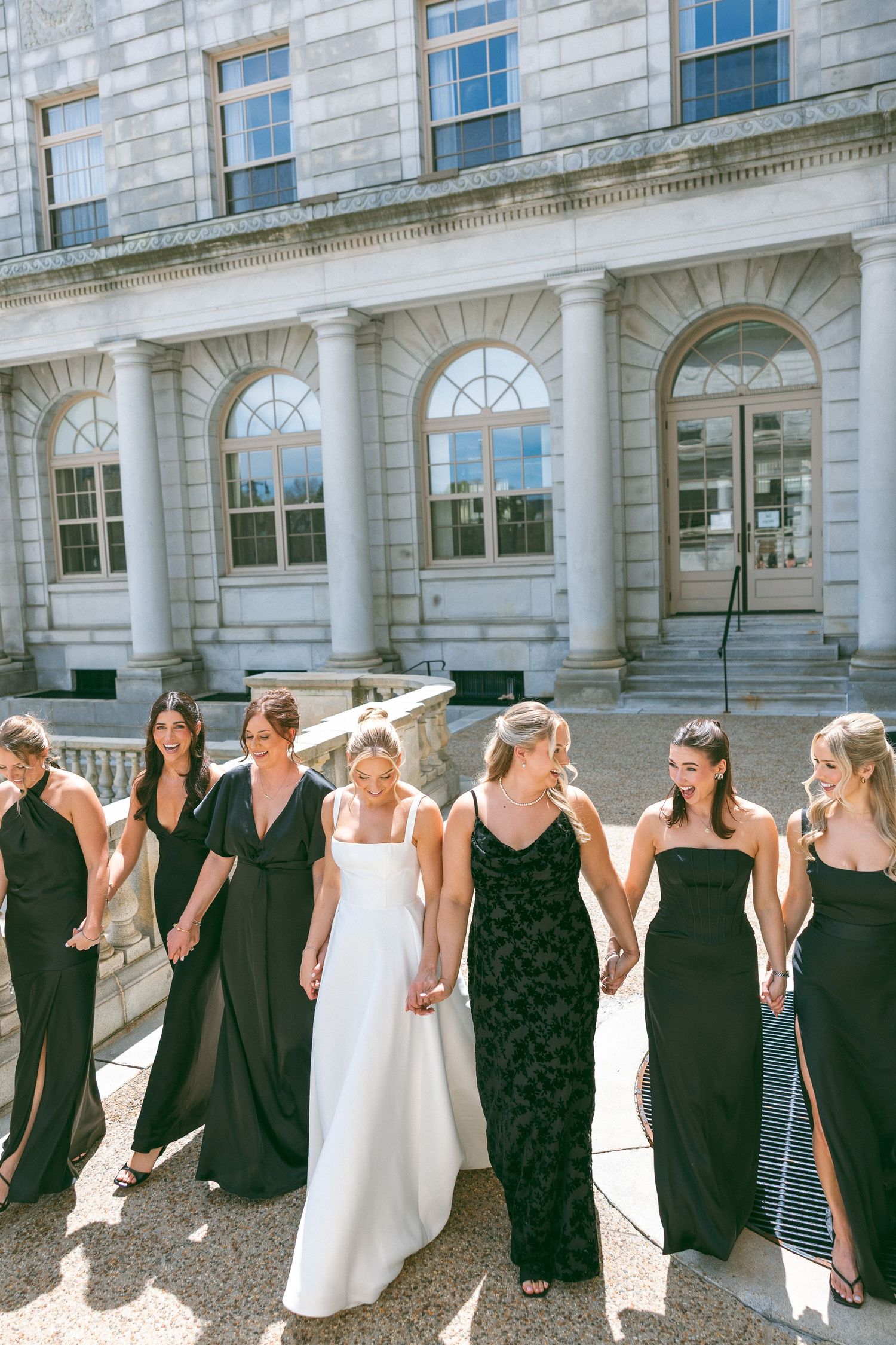 Bride in white gown walks with bridesmaids in black dresses outside a grand stone building on a sunny day.