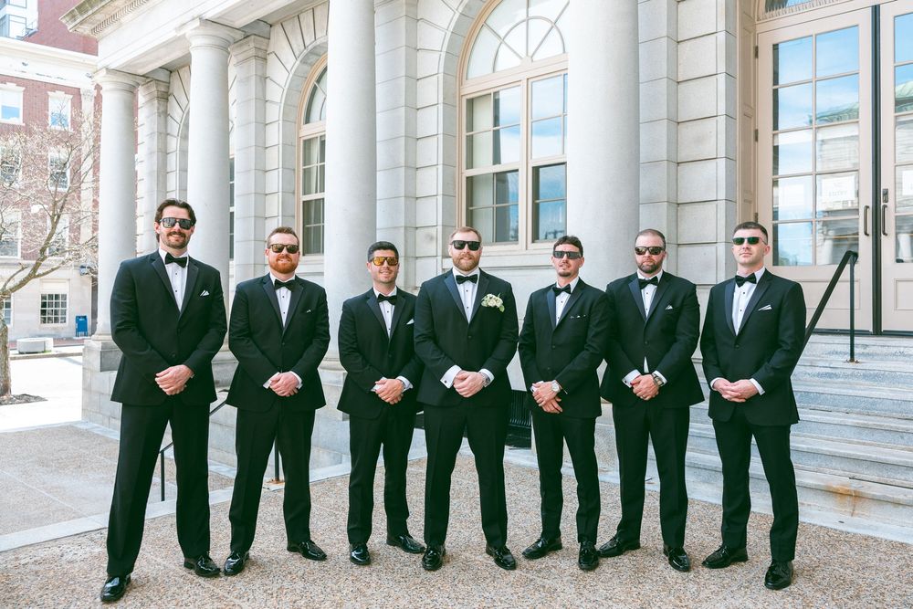 Seven groomsmen in black tuxedos and bow ties pose outside a columned building for a wedding photo.