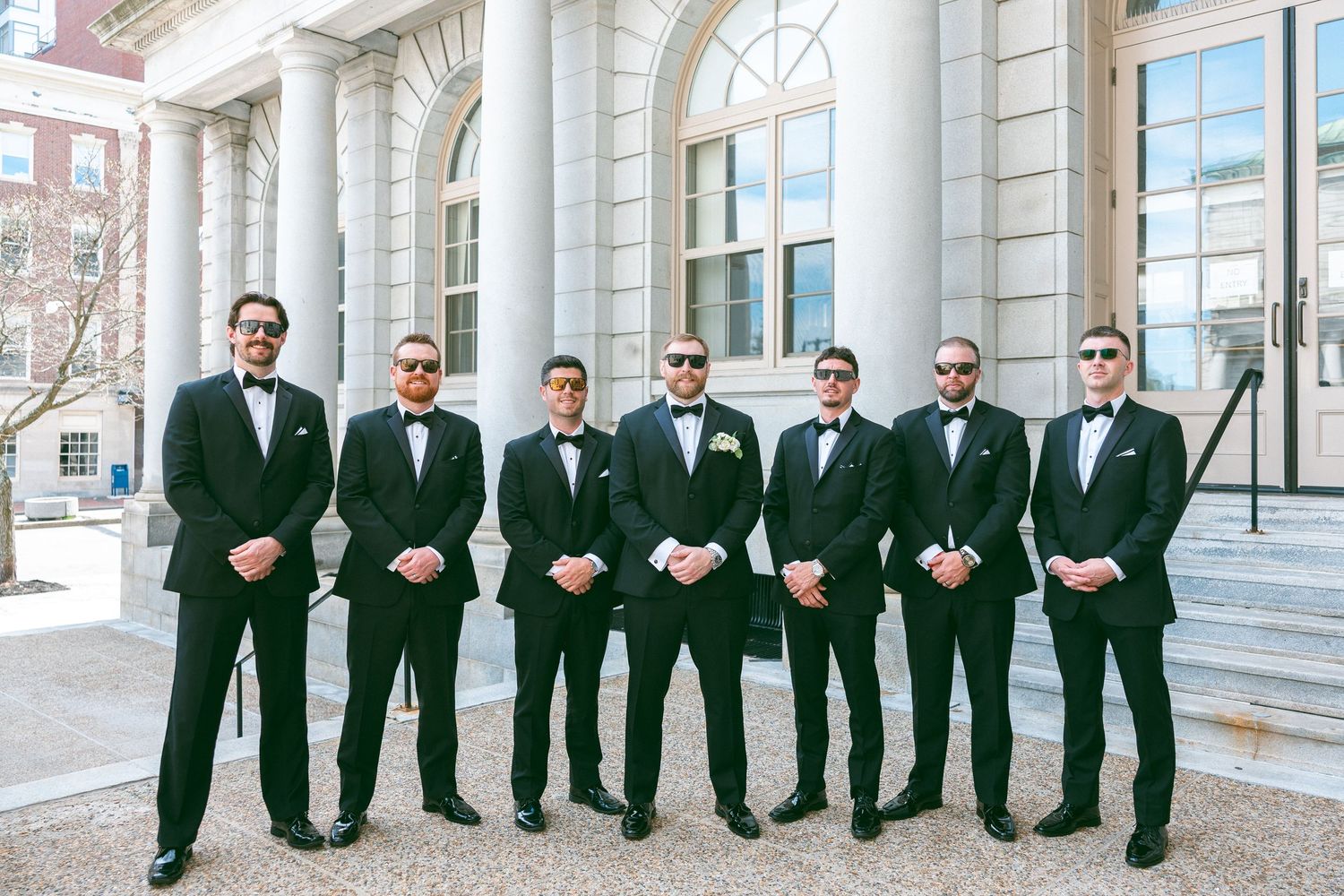 Seven groomsmen in black tuxedos and bow ties pose outside a columned building for a wedding photo.