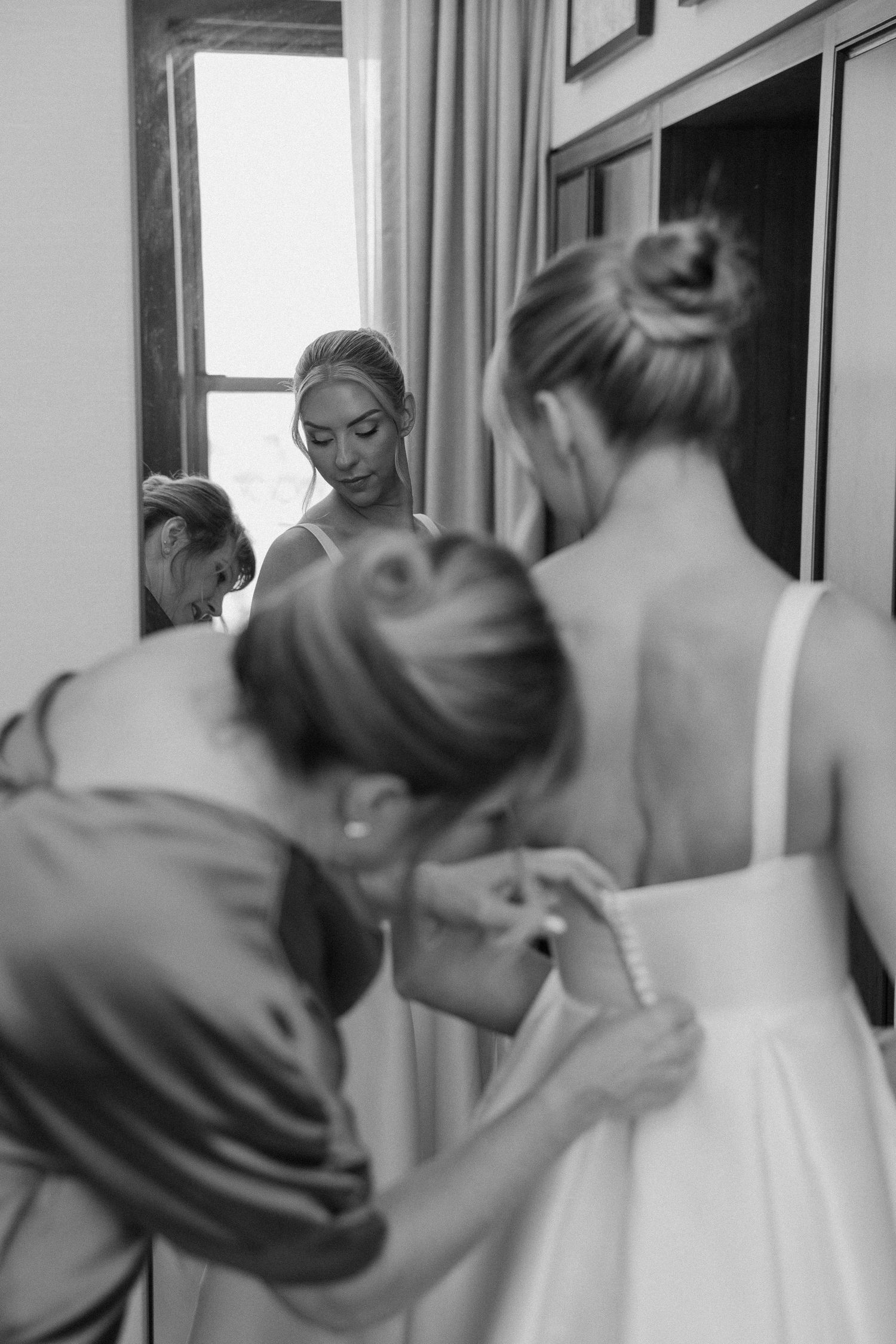 Black and white photo of bride getting wedding dress fastened by bridesmaid, with reflection in mirror.