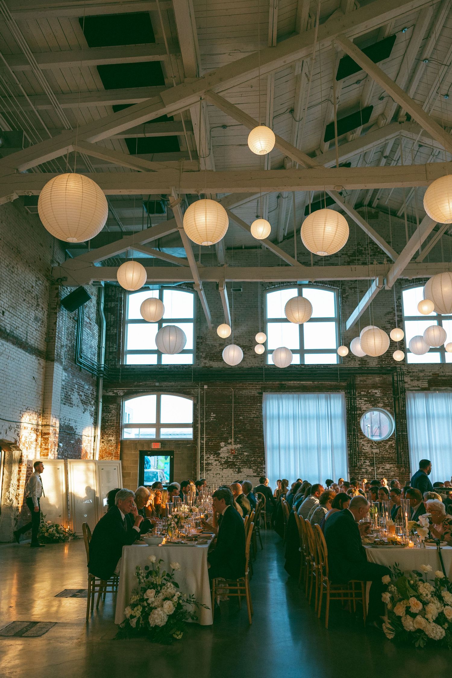 Elegant wedding reception in industrial venue with exposed brick, white paper lantern ceiling decor, and guests dining at candlelit tables.