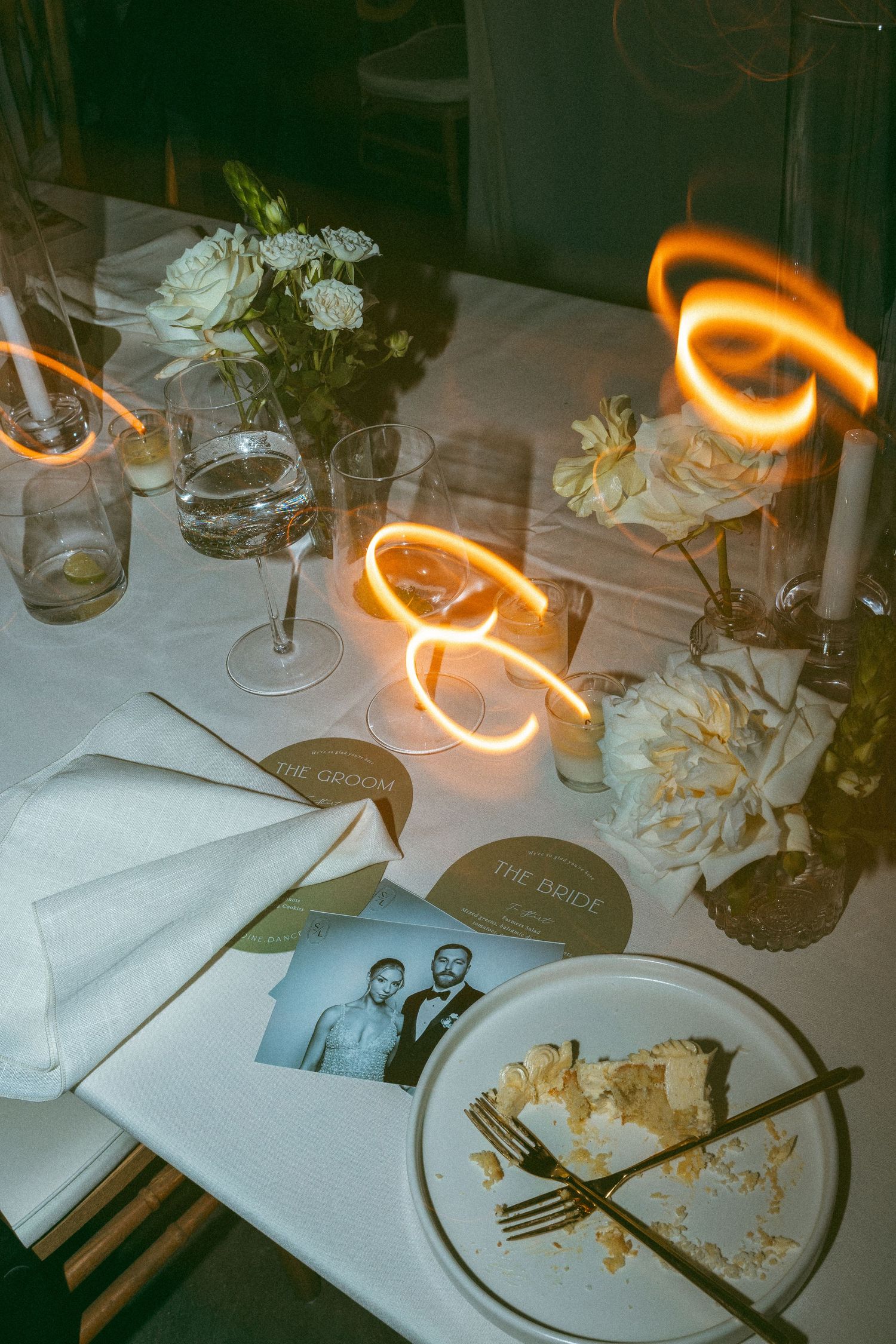 Wedding reception table with cake slice, bride and groom place cards, white flowers, candles, and light painting photography.