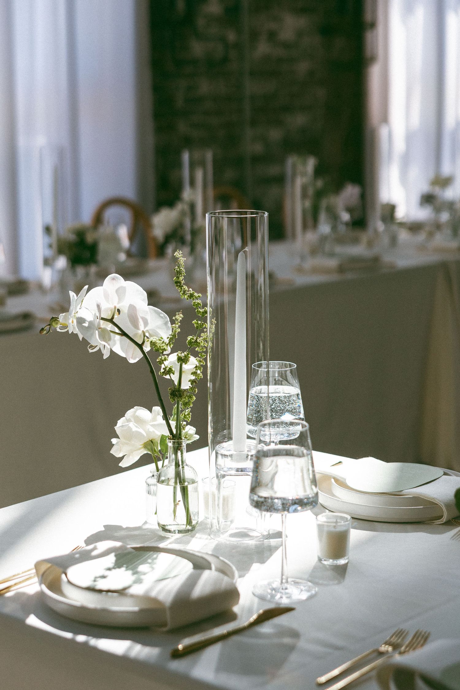 Elegant white wedding table setting with orchids, glass vases, gold cutlery, and candles in a sunlit venue.