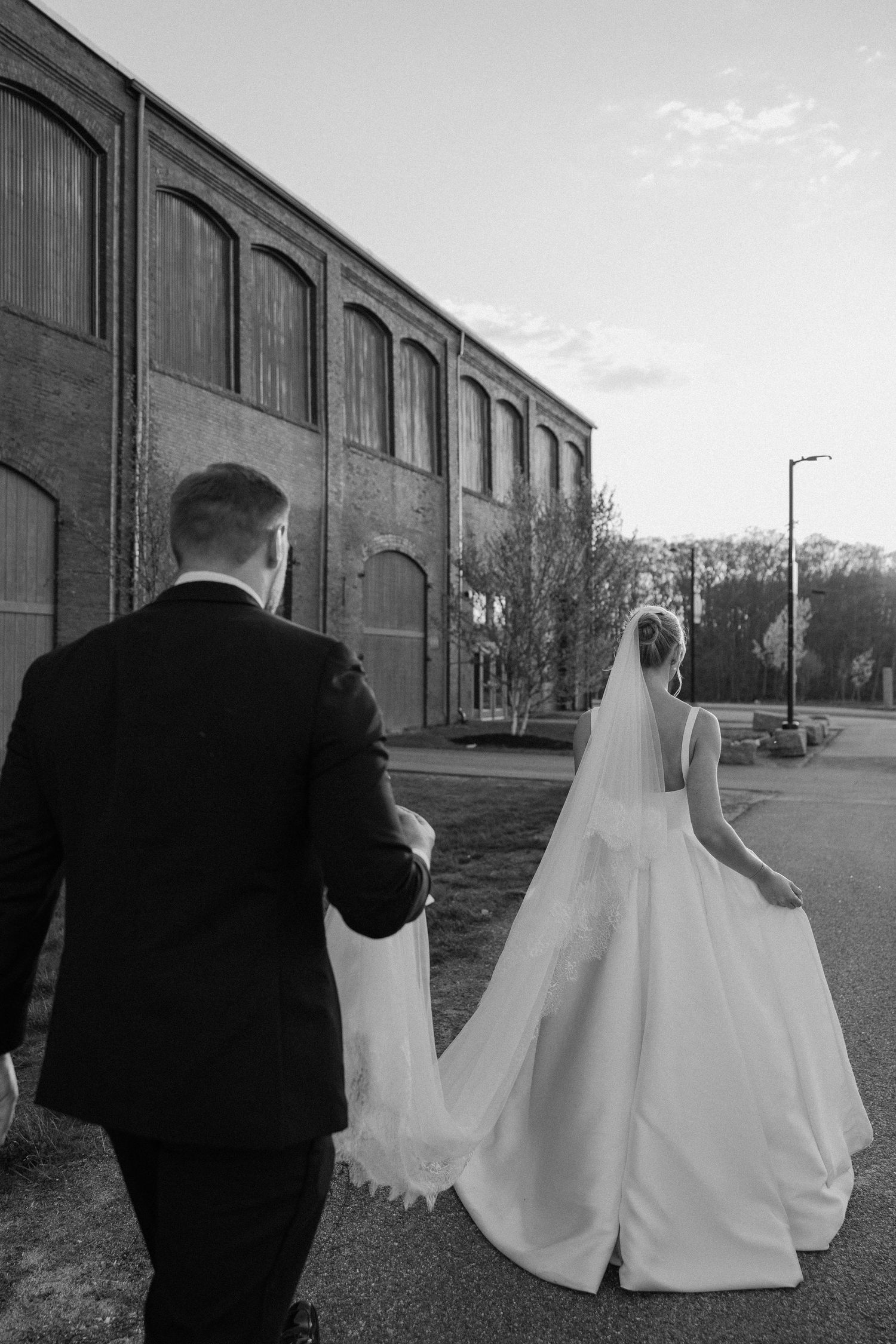 Black and white photo of a bride and groom walking hand in hand toward a rustic brick building on their wedding day.