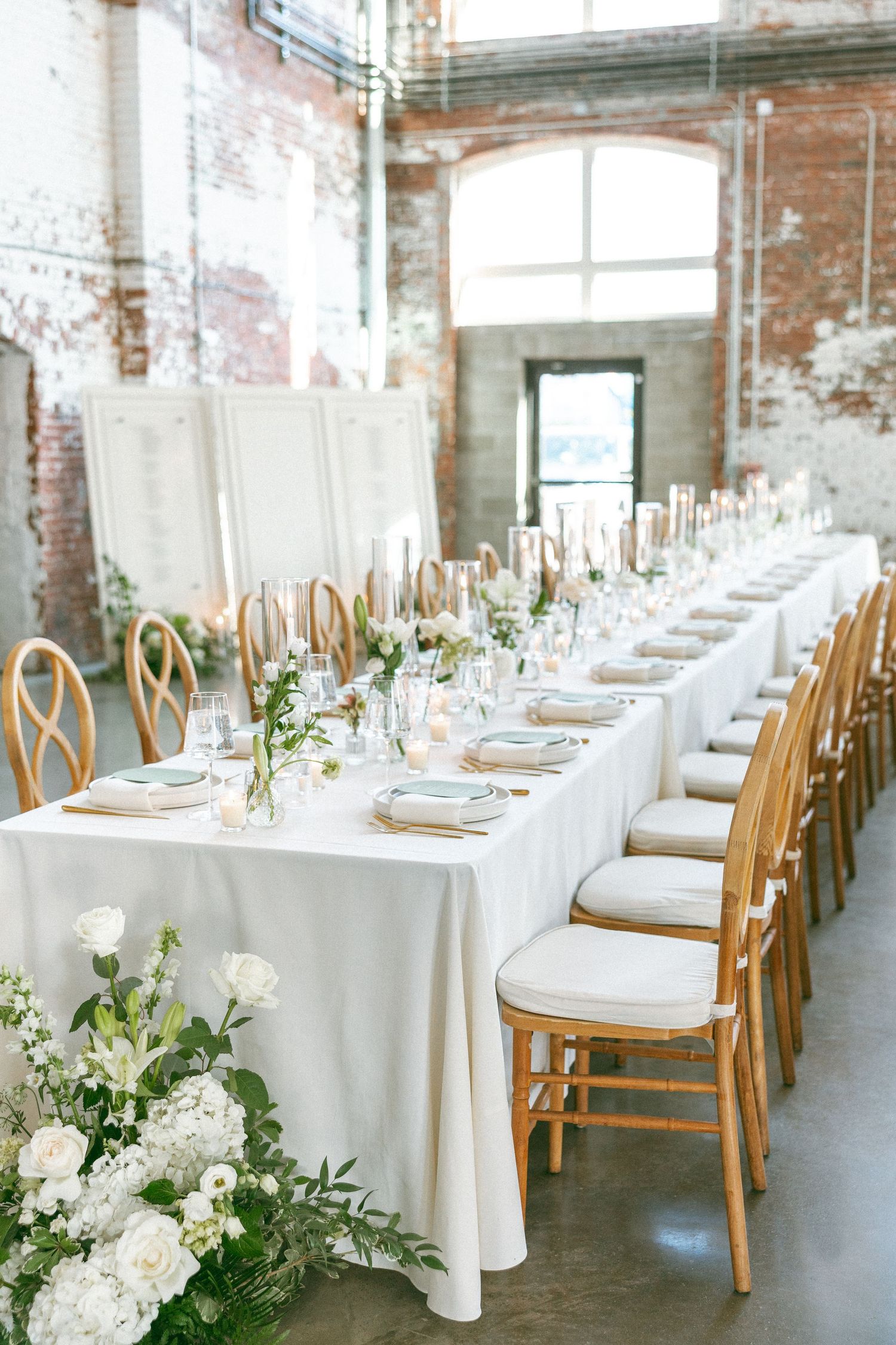 Elegant wedding reception with long white linen table, gold Chiavari chairs, white floral arrangements, and exposed brick walls.