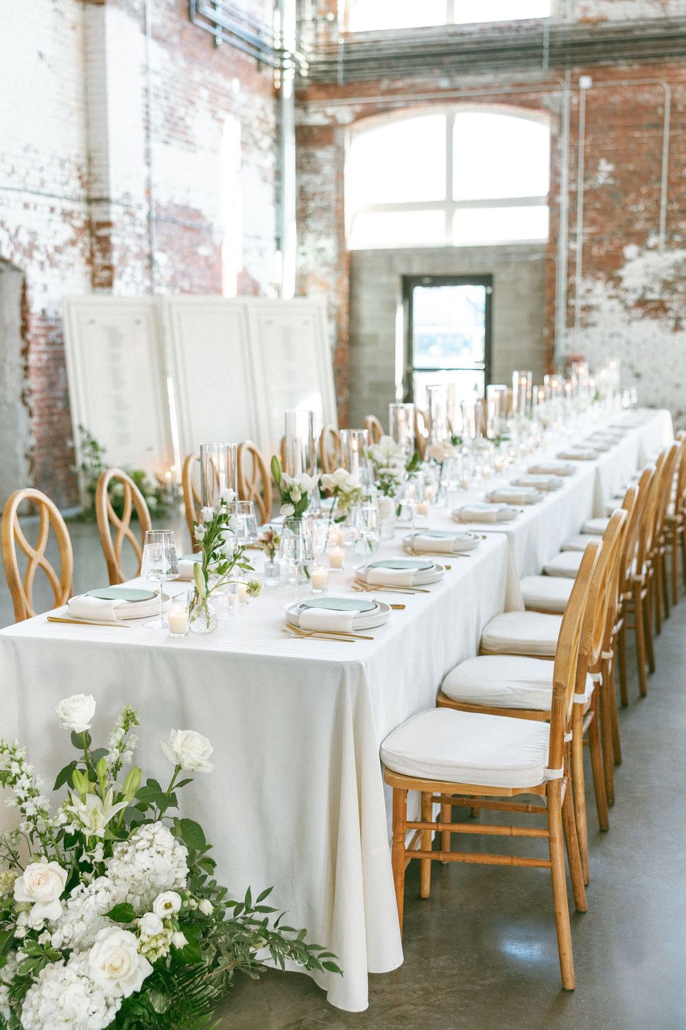 Elegant wedding reception with long white linen table, gold Chiavari chairs, white floral arrangements, and exposed brick walls.