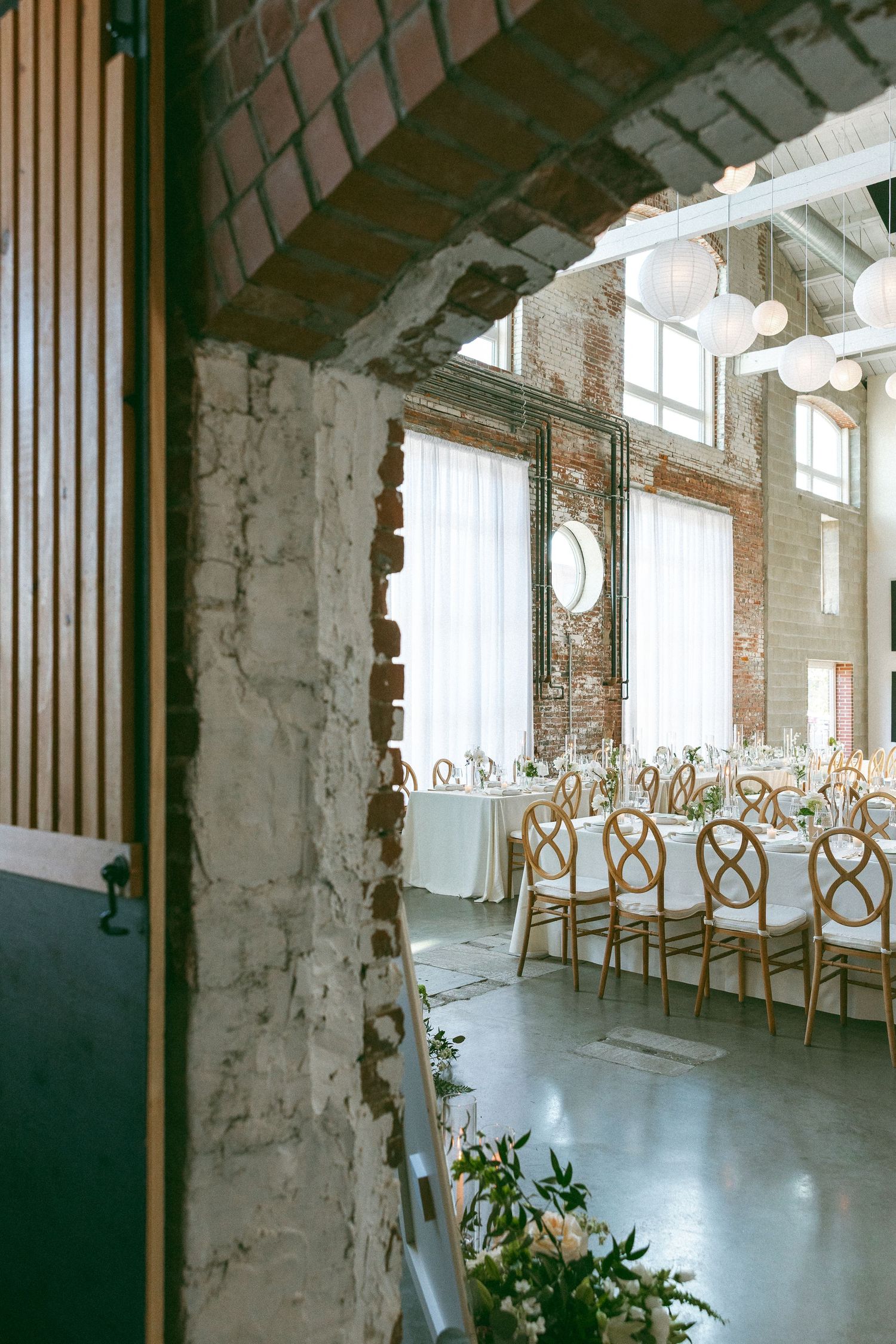 Elegant wedding reception in a rustic brick venue with long banquet tables, gold cross-back chairs, and white paper lanterns.