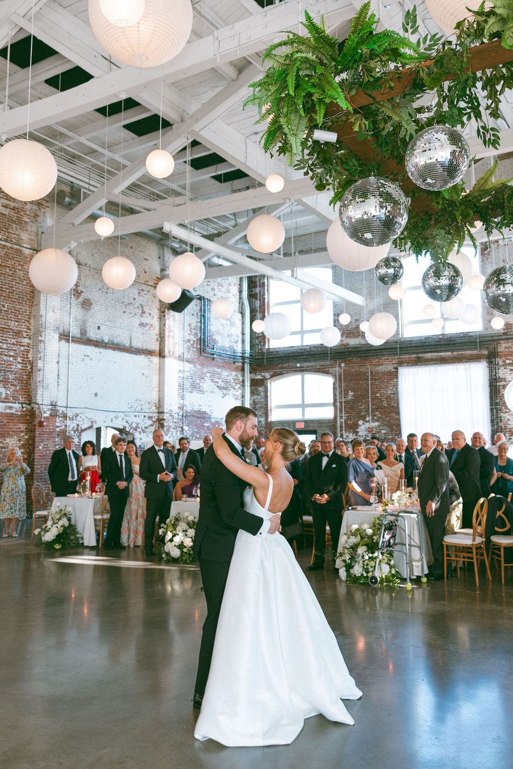 Bride and groom share first dance under disco balls and white lanterns in an industrial wedding venue.