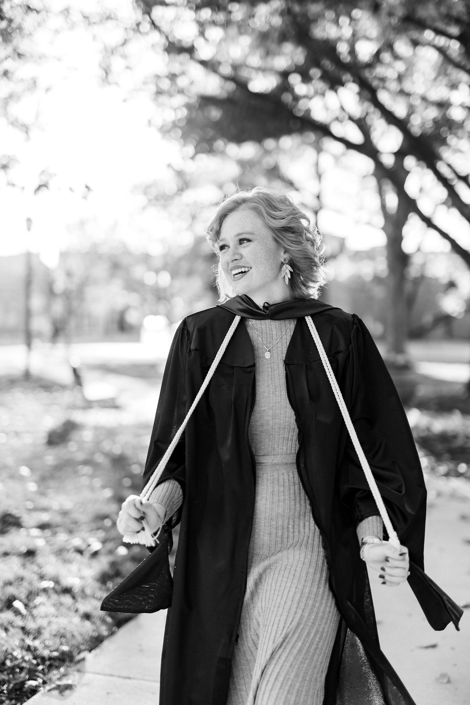 Black and white photo of a smiling woman in graduation gown walking outdoors holding cords.