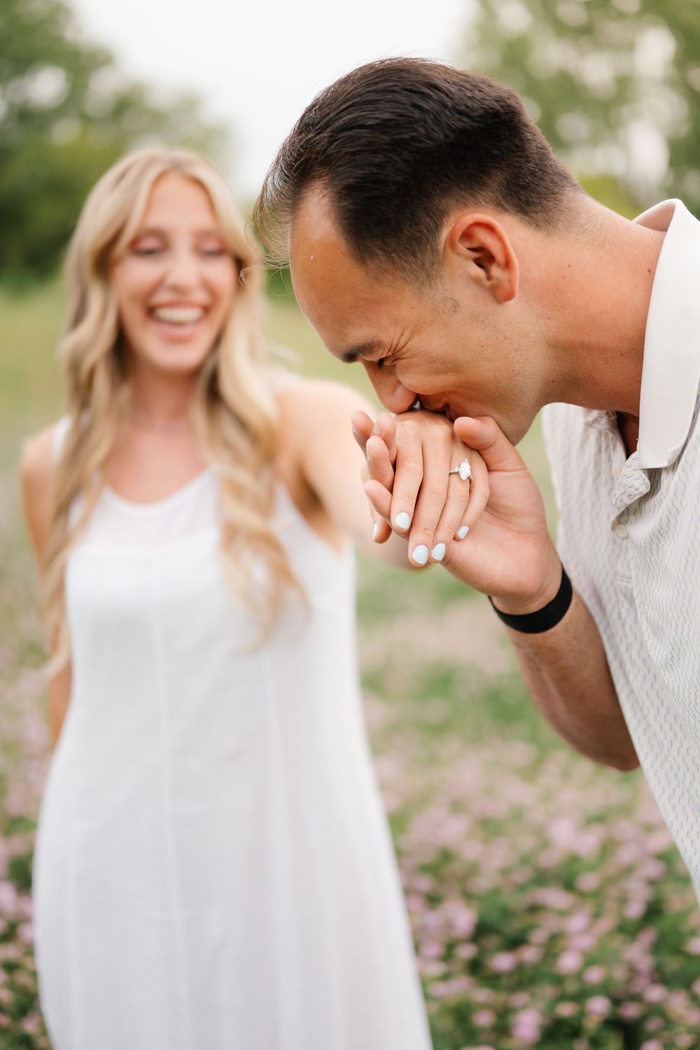 Man kissing woman's hand with engagement ring outdoors in flower field, smiling blonde bride in white dress.