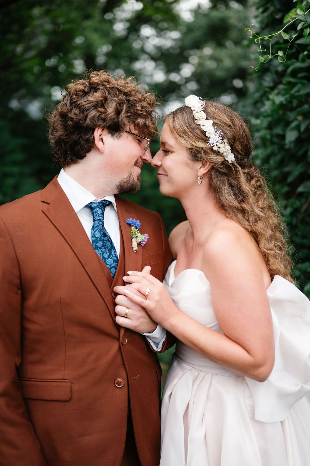 A bride and groom share a romantic nose-to-nose moment, she in a white gown with floral headpiece, he in a rust brown suit.