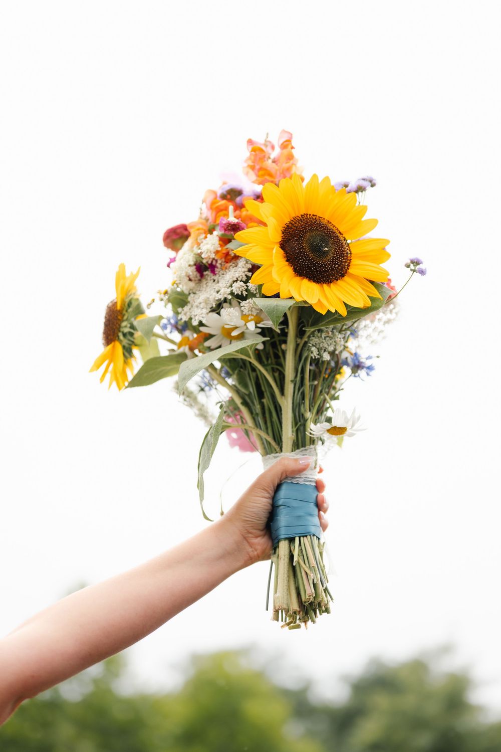 Hand holding a vibrant wildflower bouquet with sunflowers and mixed blooms wrapped in blue ribbon against sky.
