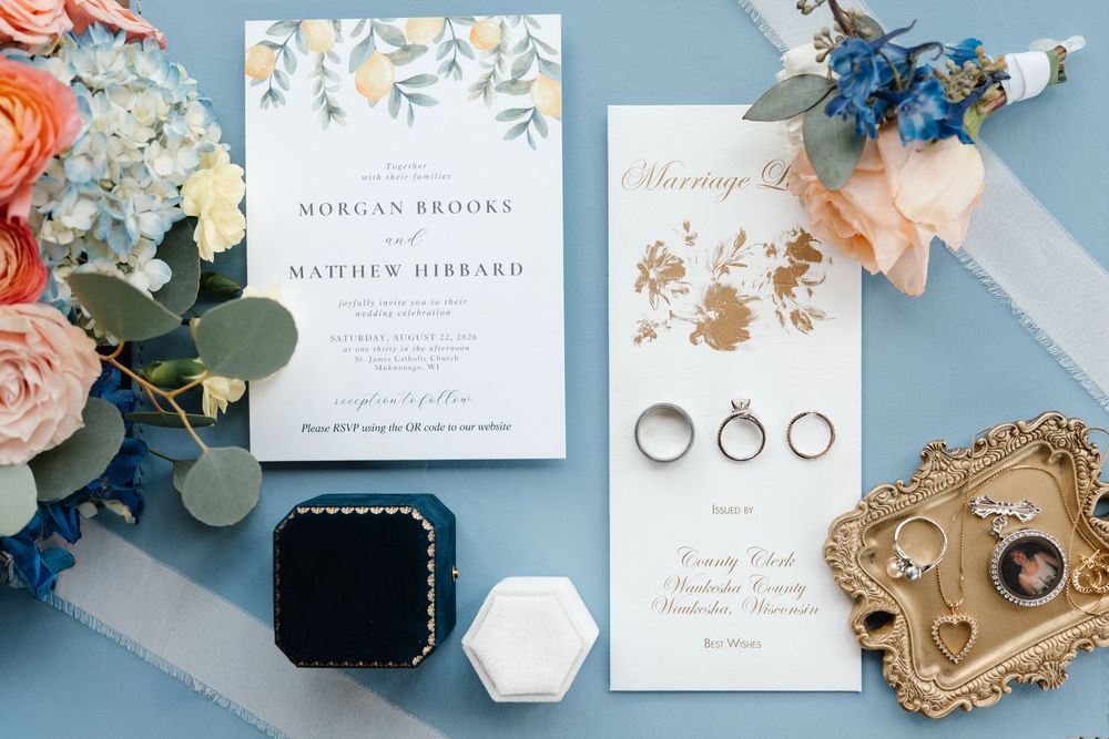 Elegant wedding flat lay with floral invitation suite, gold rings, and blue velvet ring box on a soft blue background.