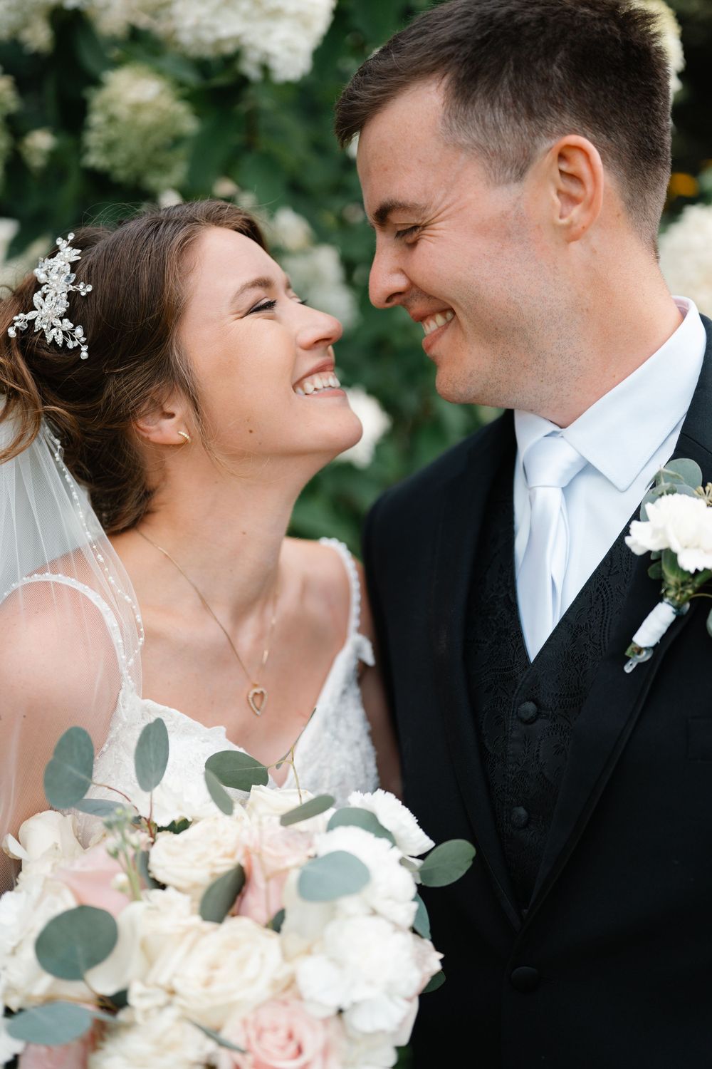 Smiling bride and groom share a joyful moment, bride holding white and blush bouquet with eucalyptus.