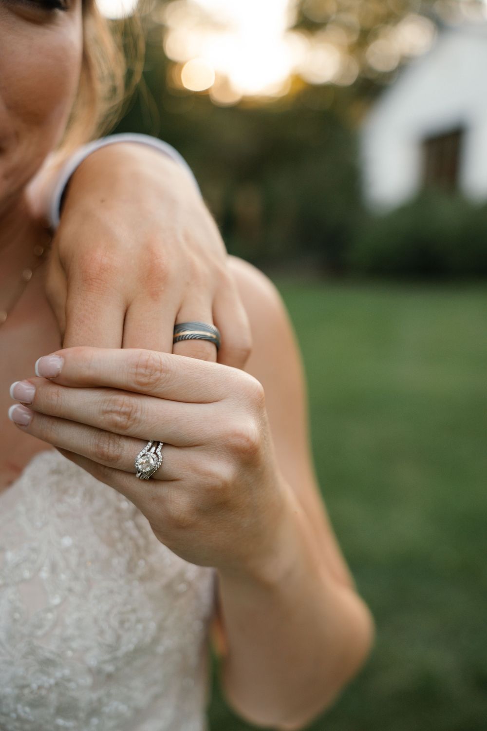 Bride displaying diamond engagement ring and groom's black wedding band on hands, outdoors with bokeh background.