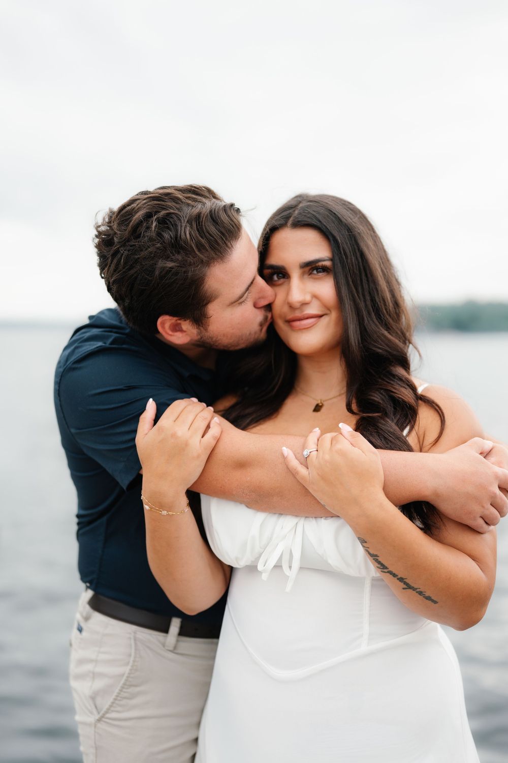 A man in a navy shirt kisses a woman in a white dress on the cheek near a lake.