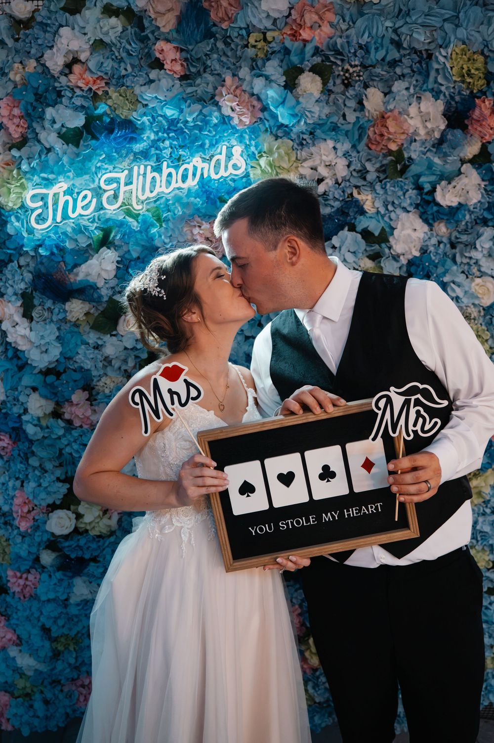 Newlyweds Mr. and Mrs. Hibbard share a kiss holding a You Stole My Heart sign in front of a floral wall.