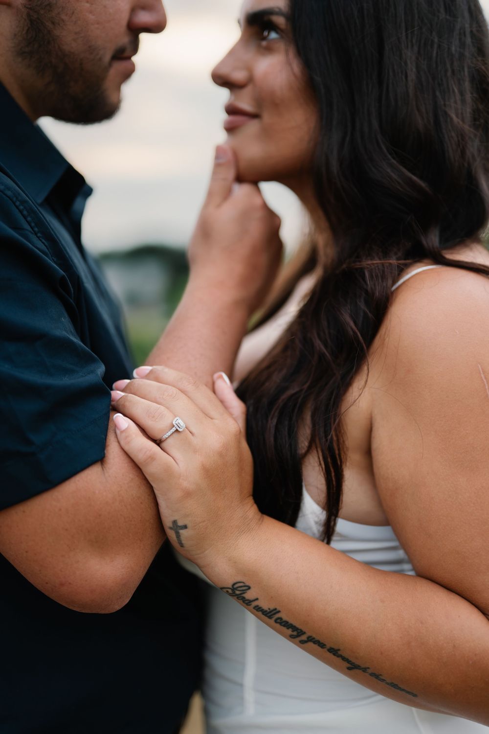 Couple in close embrace showcasing engagement ring, woman in white dress with script tattoo gazing up at partner outdoors.