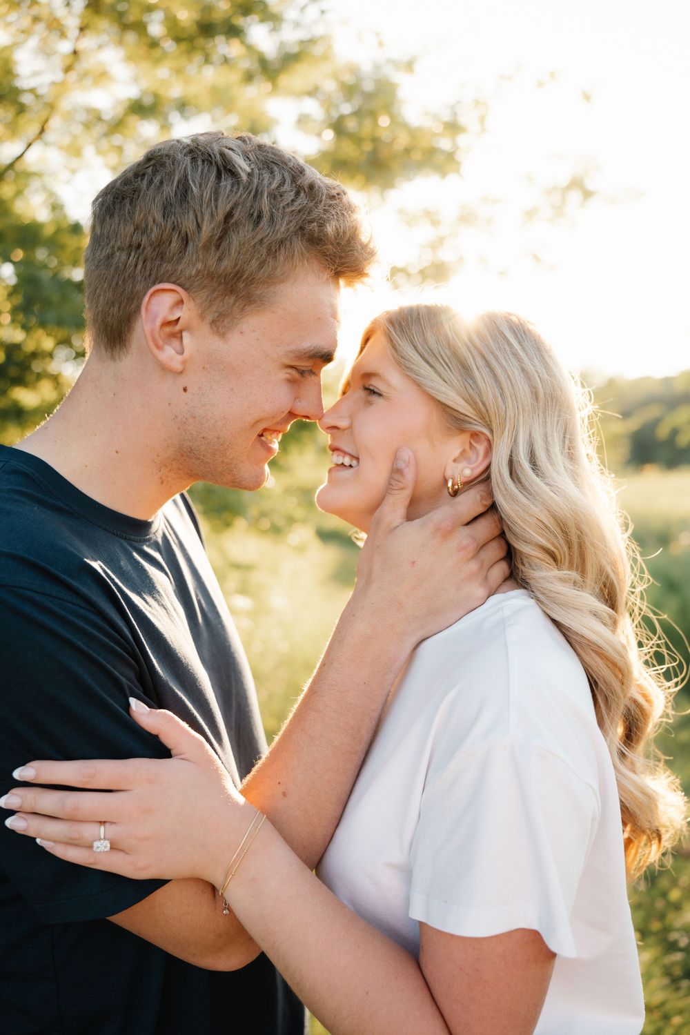 A smiling couple sharing a romantic moment outdoors, the woman wearing an engagement ring, bathed in golden sunlight.