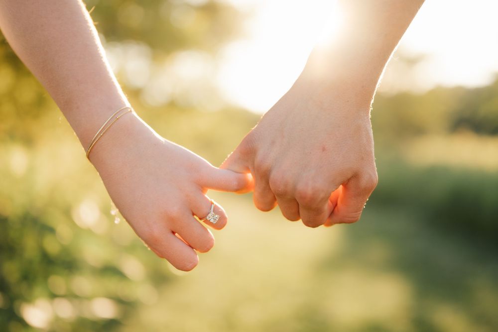 Two people holding hands with interlocked fingers outdoors in golden sunlight, one wearing a diamond engagement ring.