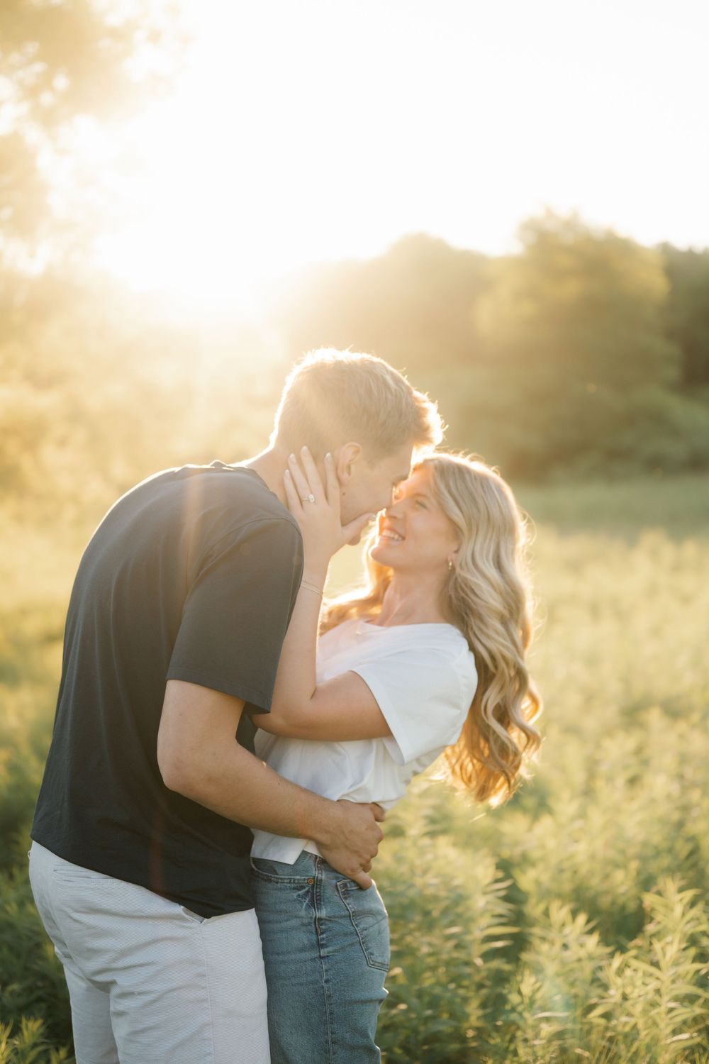 Couple sharing a romantic moment in a sunlit field during golden hour, woman smiling in white top and jeans.