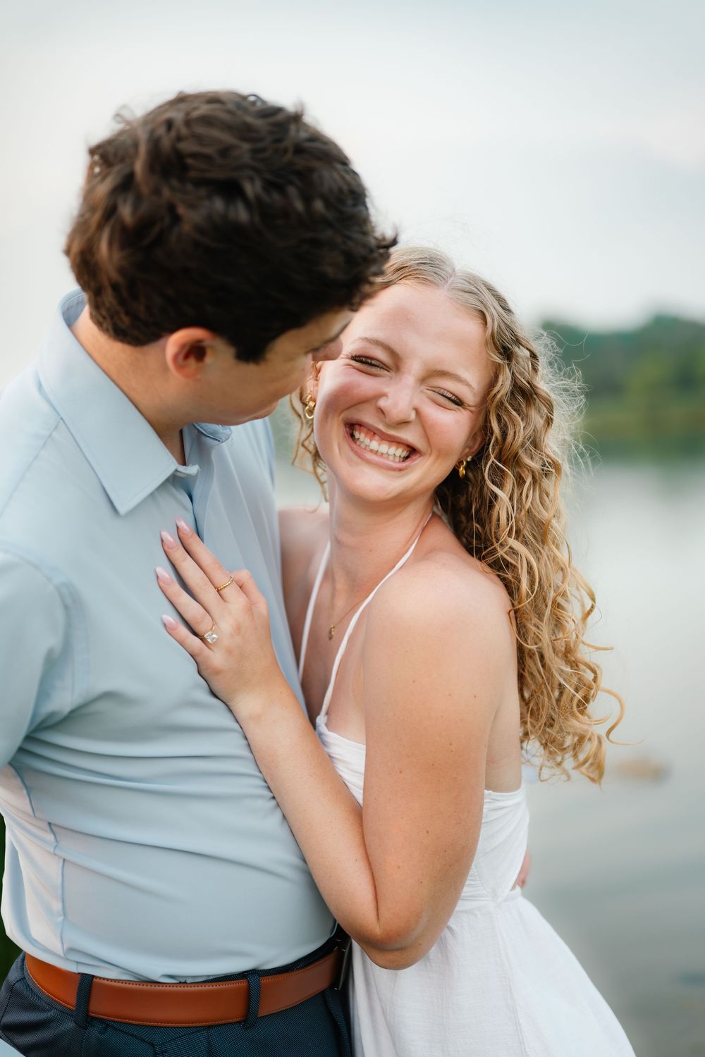 A joyful bride in white laughs as her groom in a blue shirt whispers in her ear near a lake.