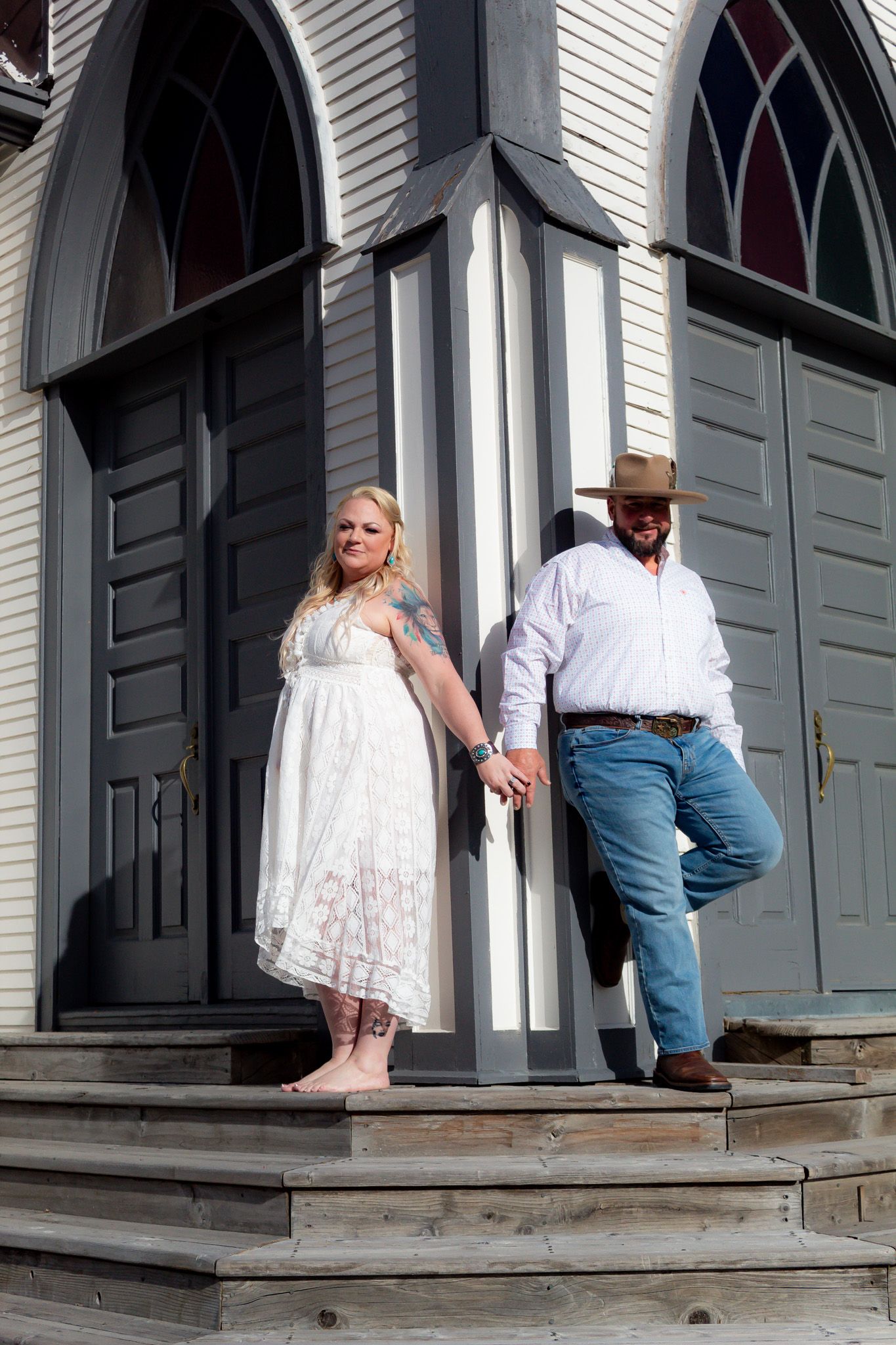 Couple posing on the steps of a white church with gothic arched windows.