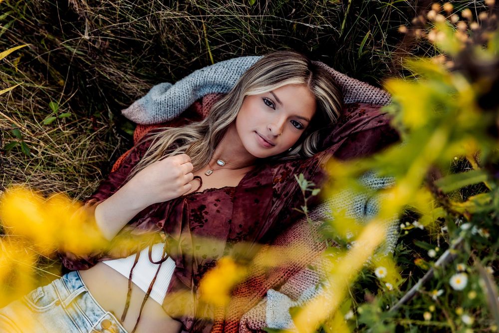 Young blonde woman lying in a field of wildflowers, wearing a burgundy dress, surrounded by yellow blooms and green grass.