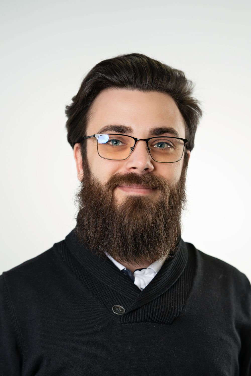Professional headshot of person with dark beard and glasses wearing a black sweater against white background.