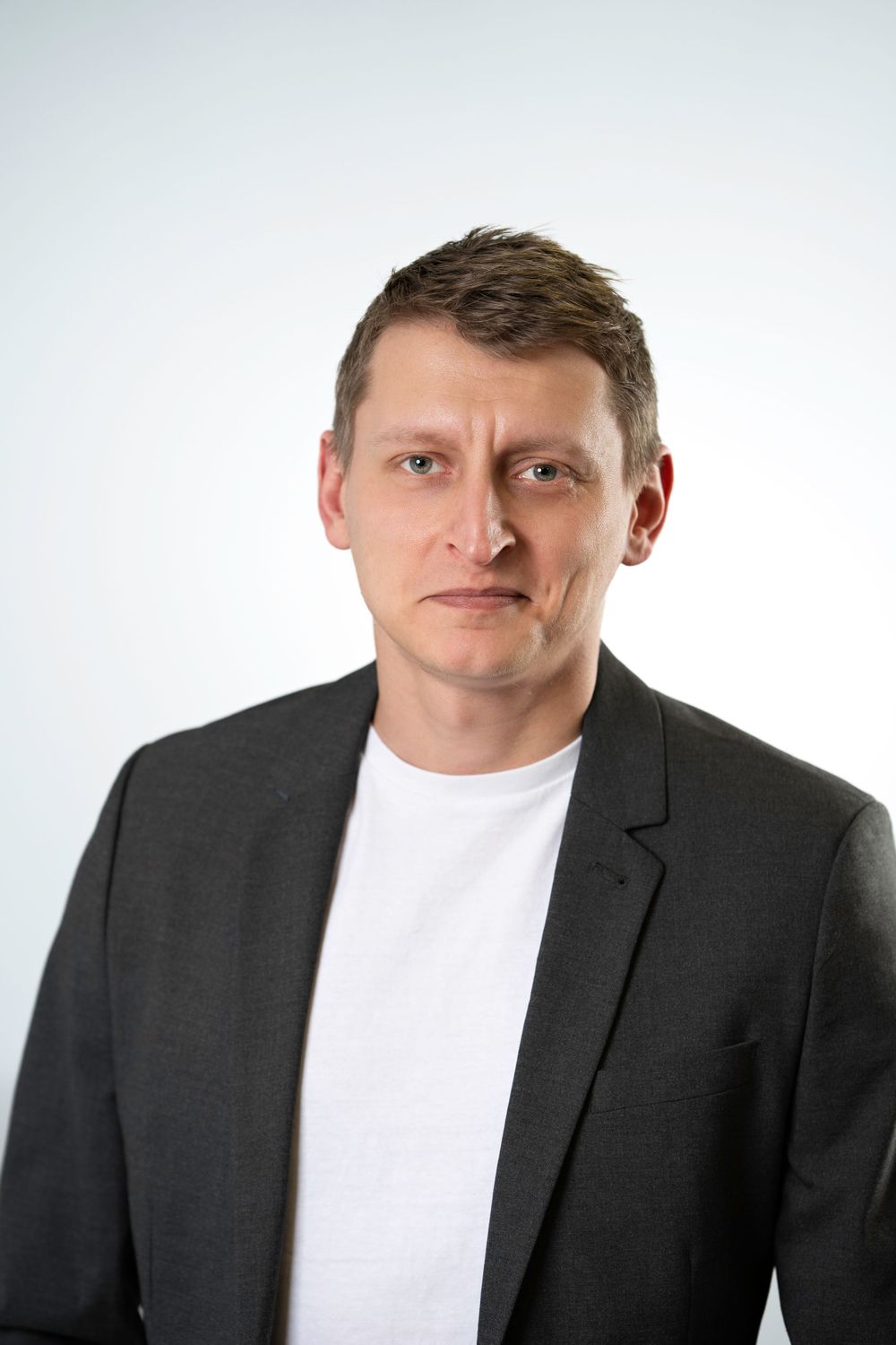 Professional headshot of person in white t-shirt and dark blazer against light background.