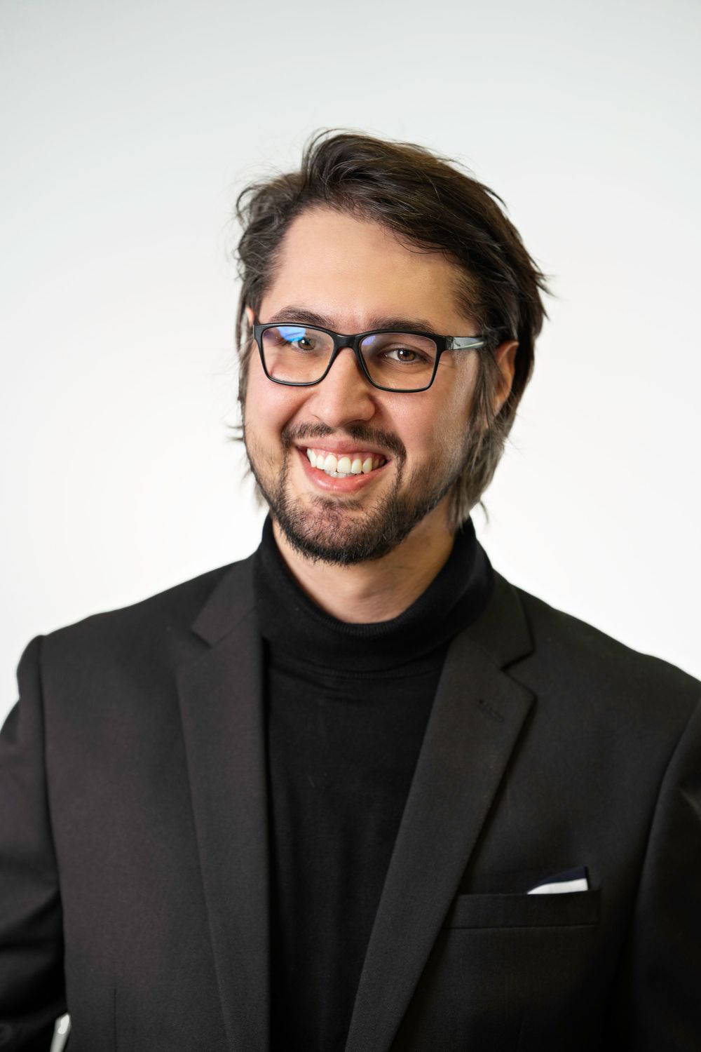 Professional portrait of person wearing glasses and black turtleneck with blazer against white backdrop.