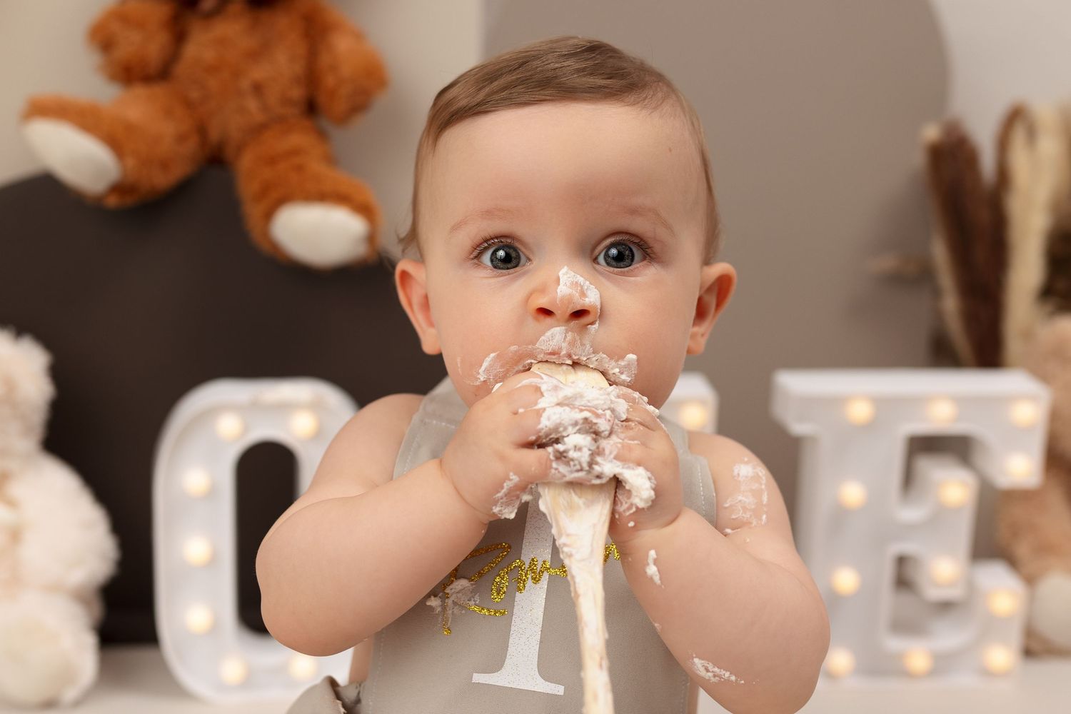 Baby enjoying a messy first birthday celebration with cake and stuffed animals in the background.