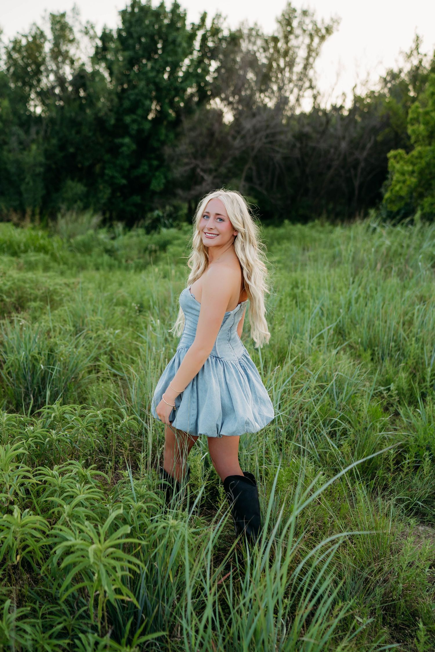 Smiling blonde woman in a light blue mini dress and black boots standing in a lush green field at dusk.