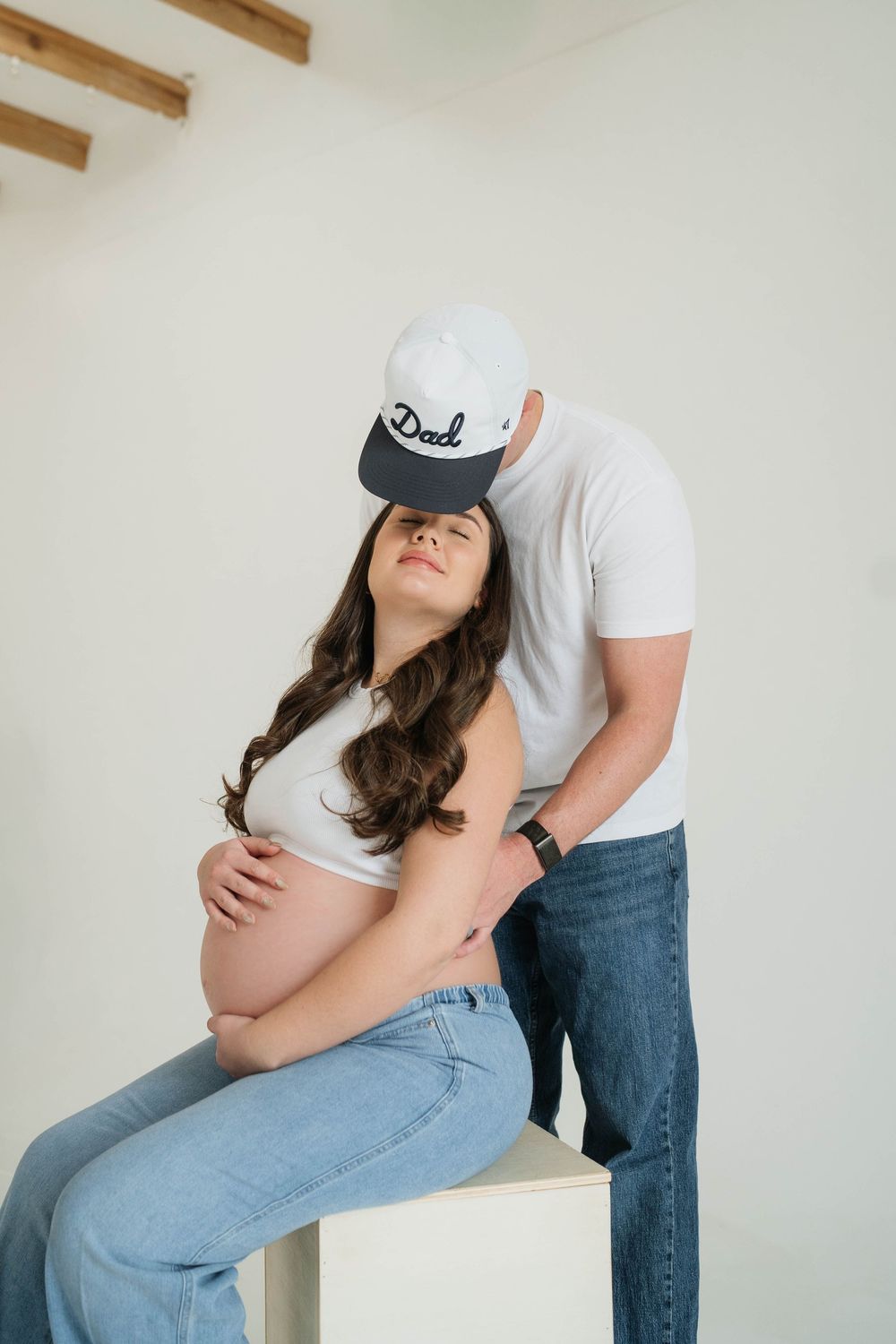 A pregnant woman in jeans and white shirt poses against a white wall in three similar artistic maternity photos.