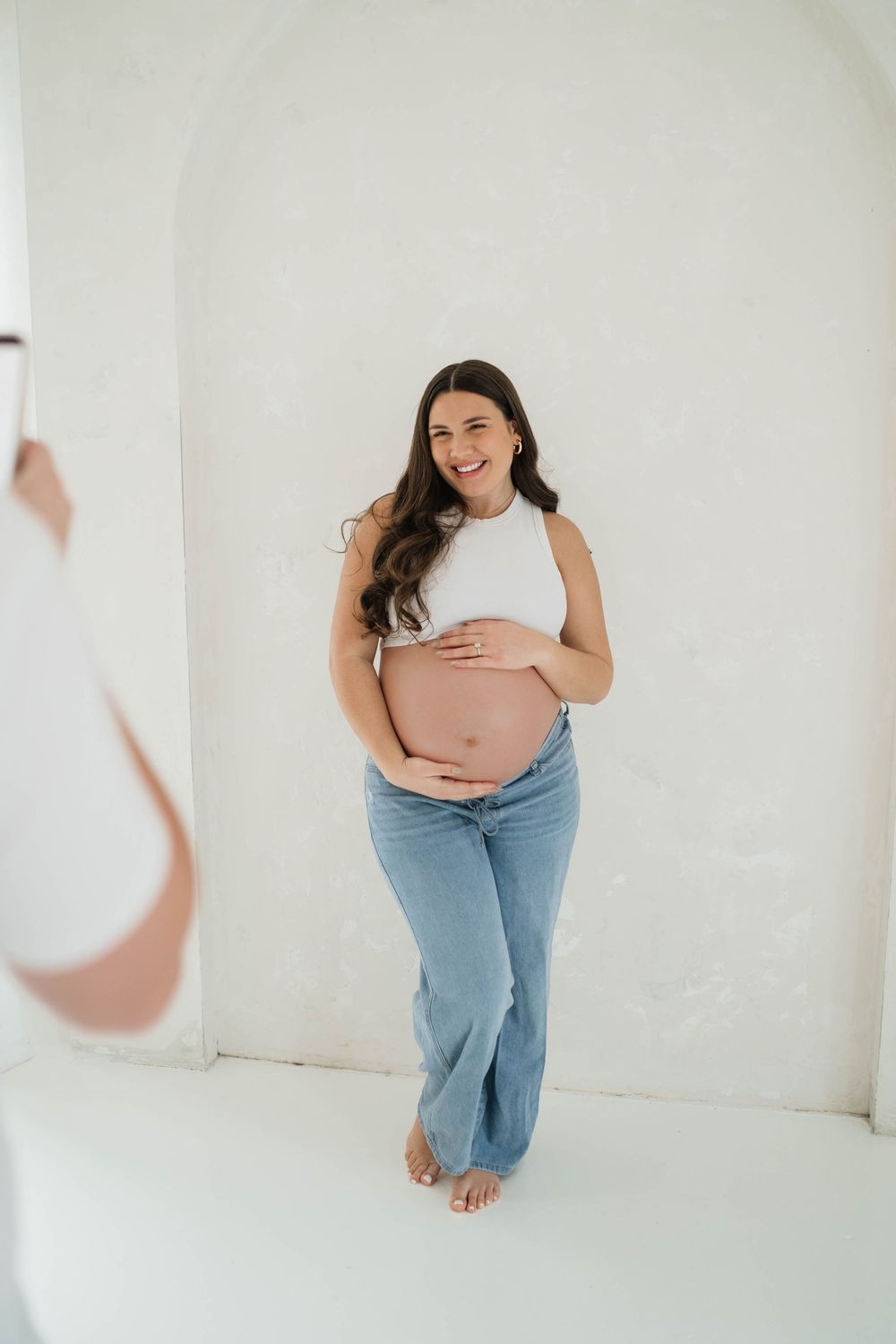 A pregnant woman in a white sleeveless top and light blue jeans stands barefoot against a white wall.