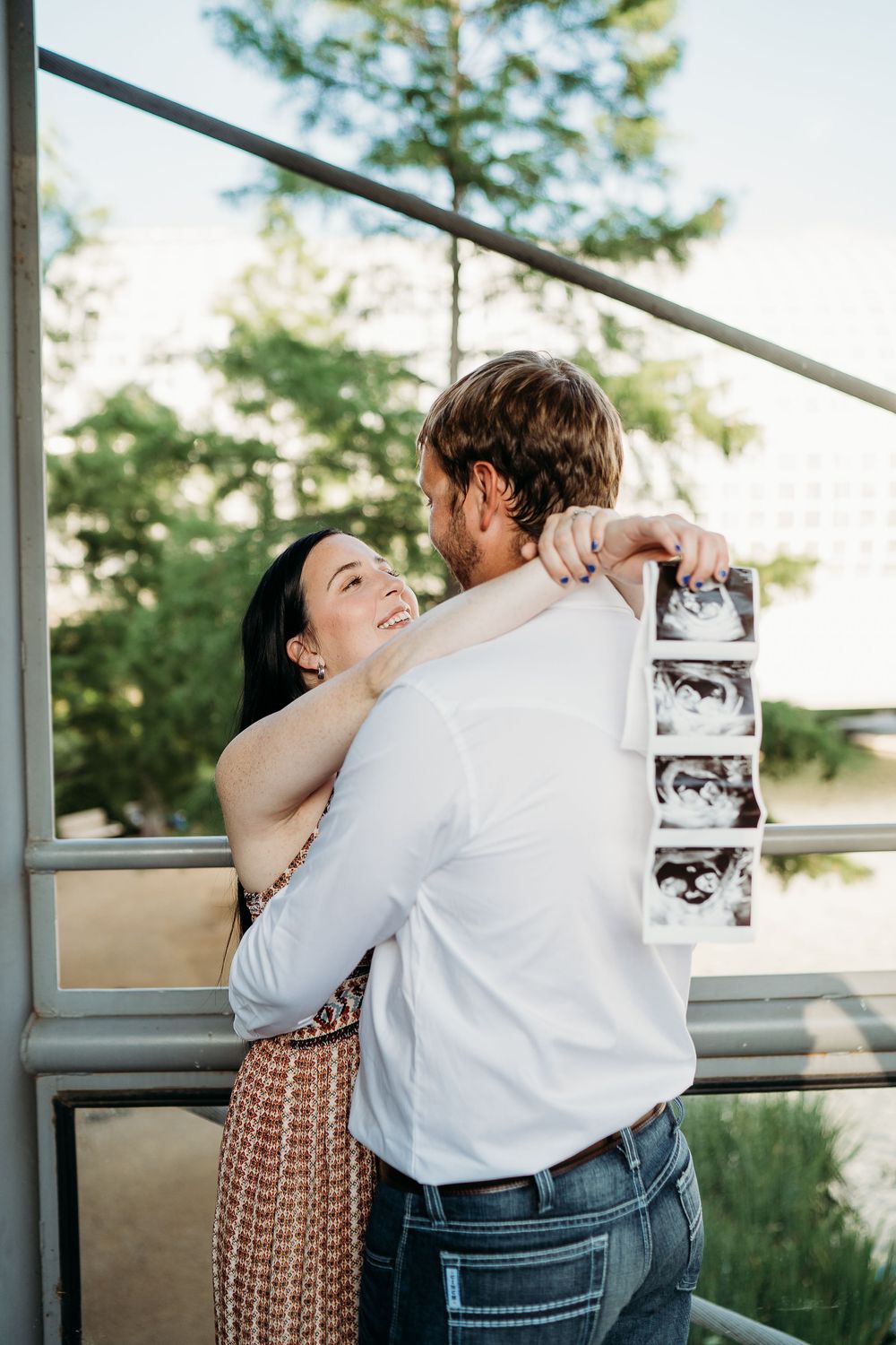 A couple embraces on a balcony while holding ultrasound photos during a pregnancy announcement photoshoot outdoors.
