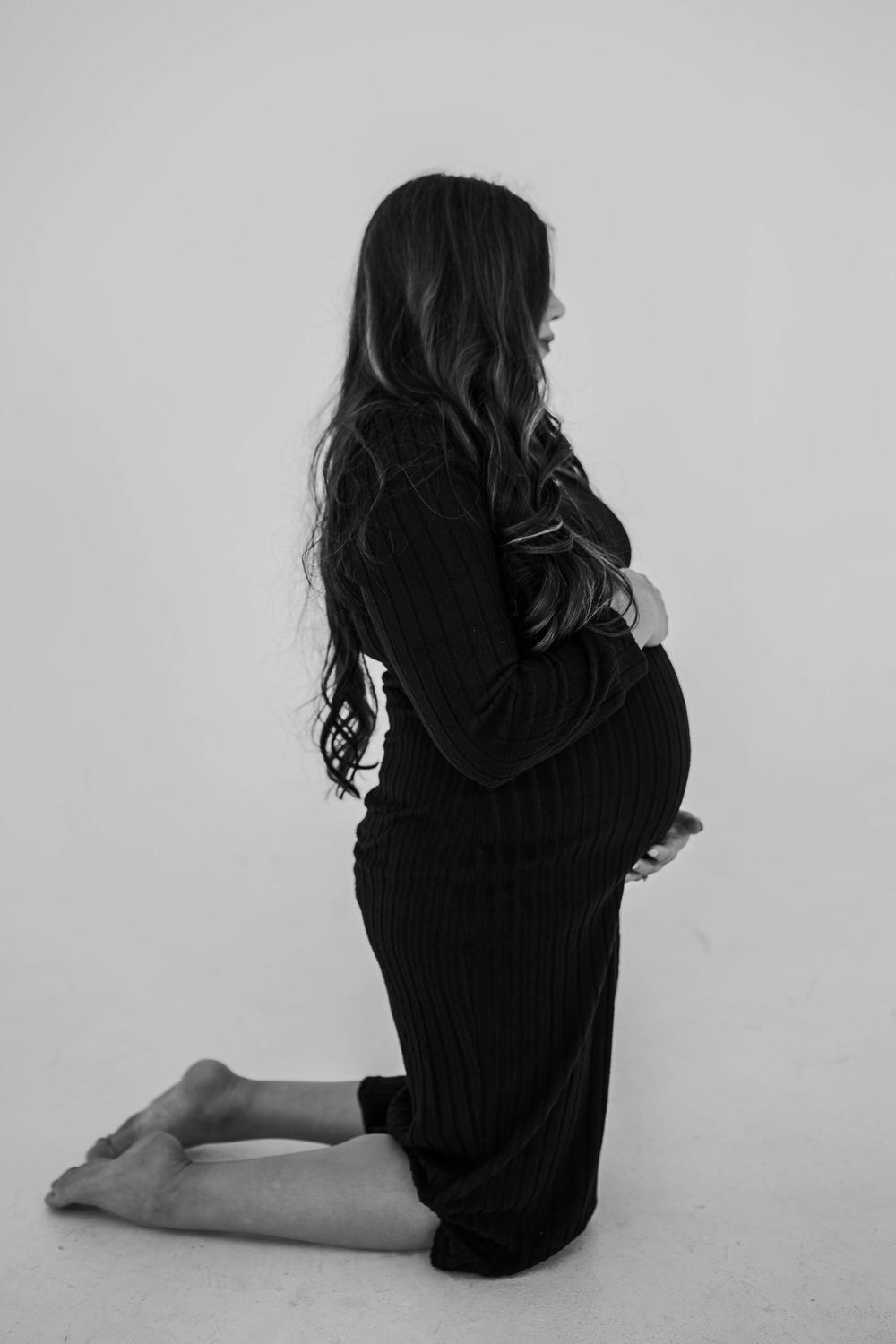 Black and white maternity photo of an expectant mother in a long dress kneeling in profile against a light background.