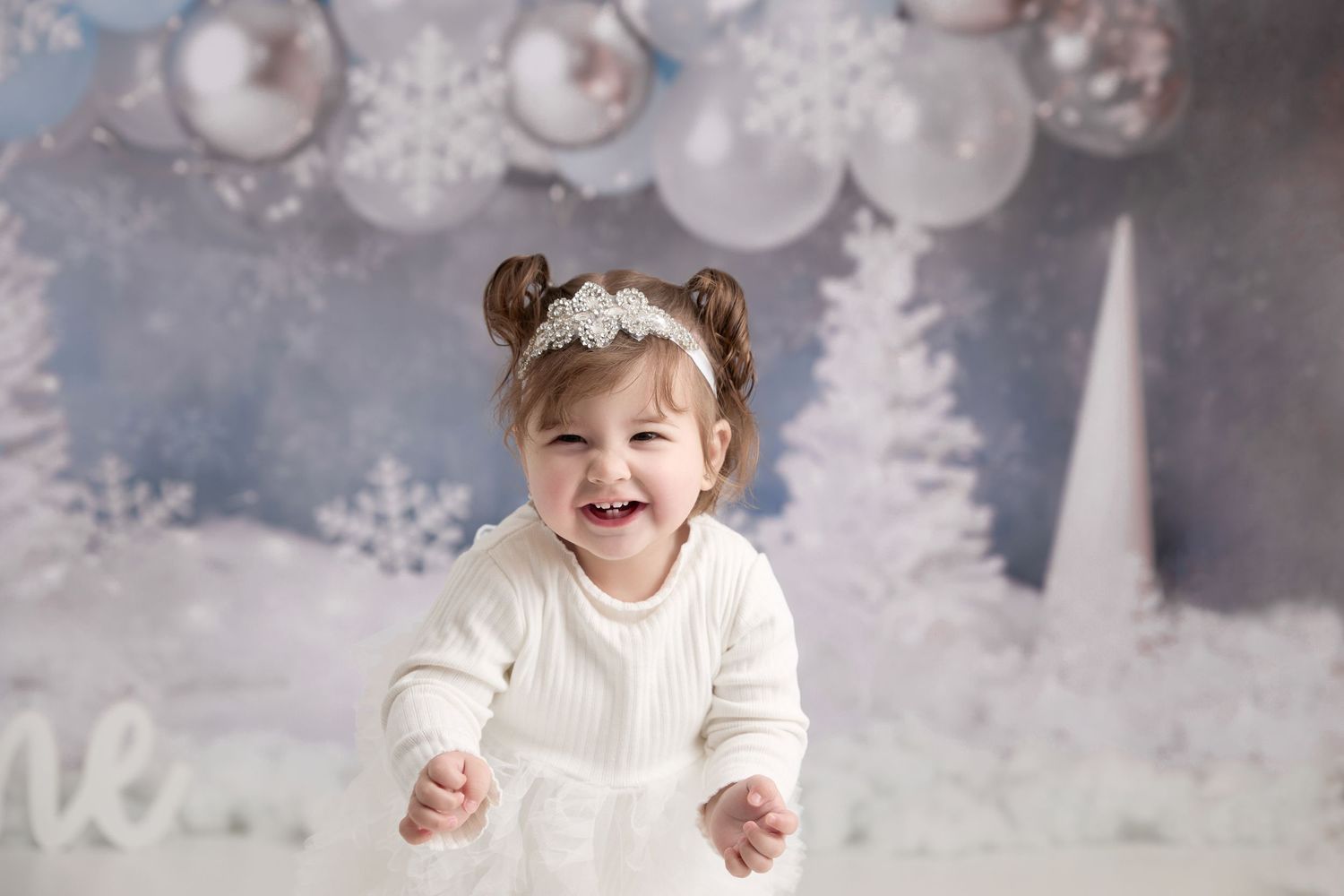 Smiling child wearing white dress and hair buns in winter wonderland holiday photo setting with white trees and ornaments. Port Chester, NY