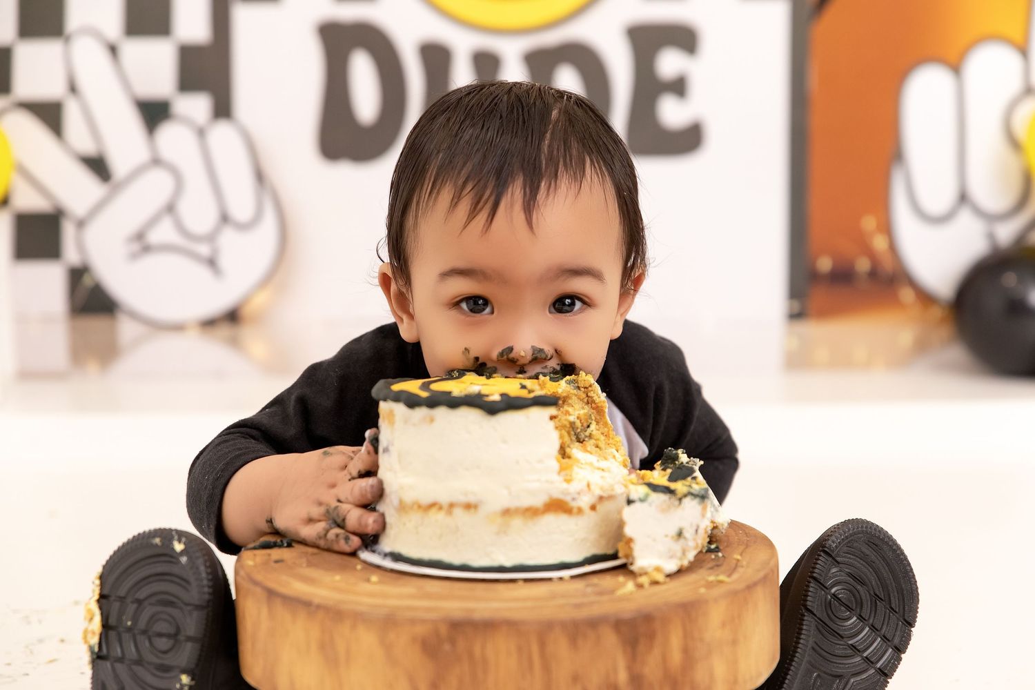 A baby boy in a black outfit smashes his face into a small birthday cake on a wooden stand at a cake smash photoshoot.